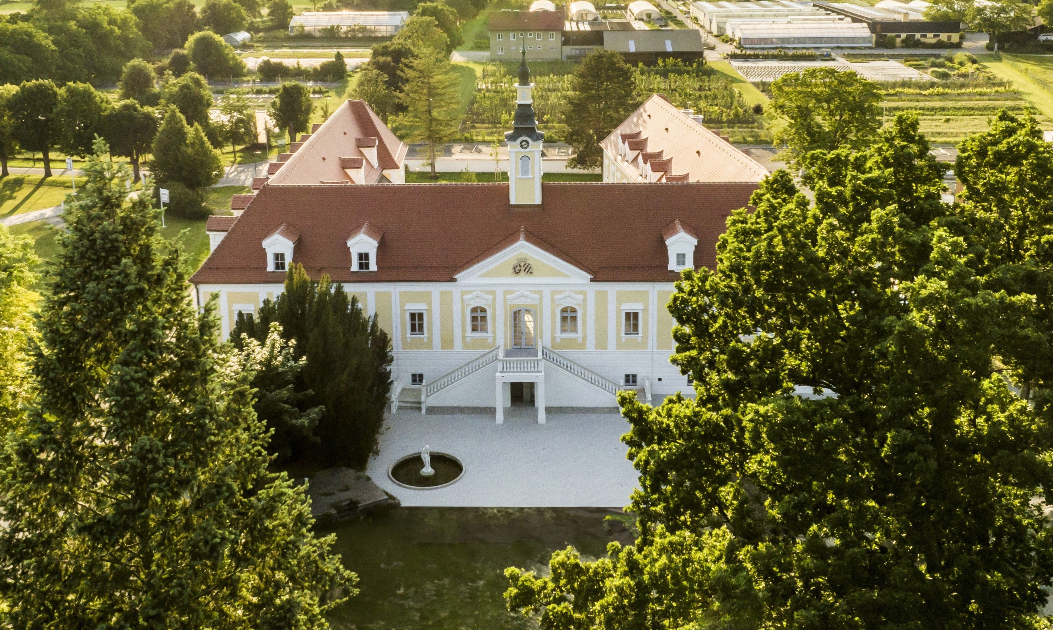 Luftaufnahme eines großen, gelben Gebäudes mit rotem Dach und Turm, umgeben von Bäumen und Grünflächen.