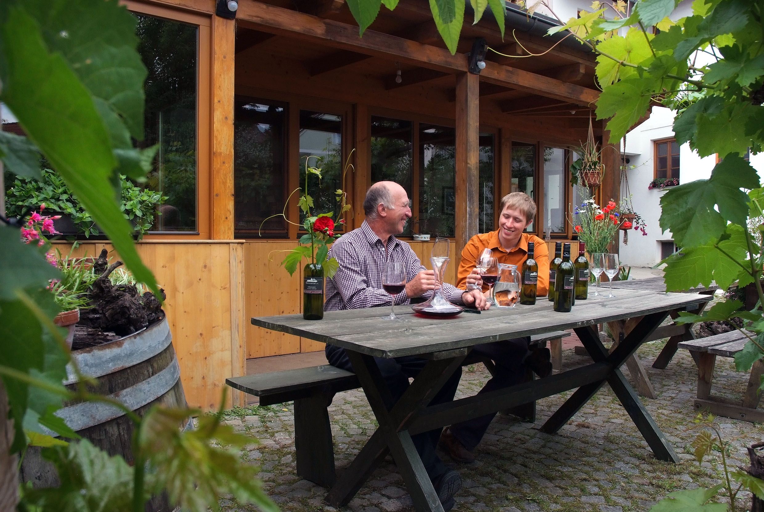 Zwei Männer sitzen an einem Holztisch im Freien und verkosten Wein. Umgeben von Pflanzen und einem Holzgebäude im Hintergrund.
