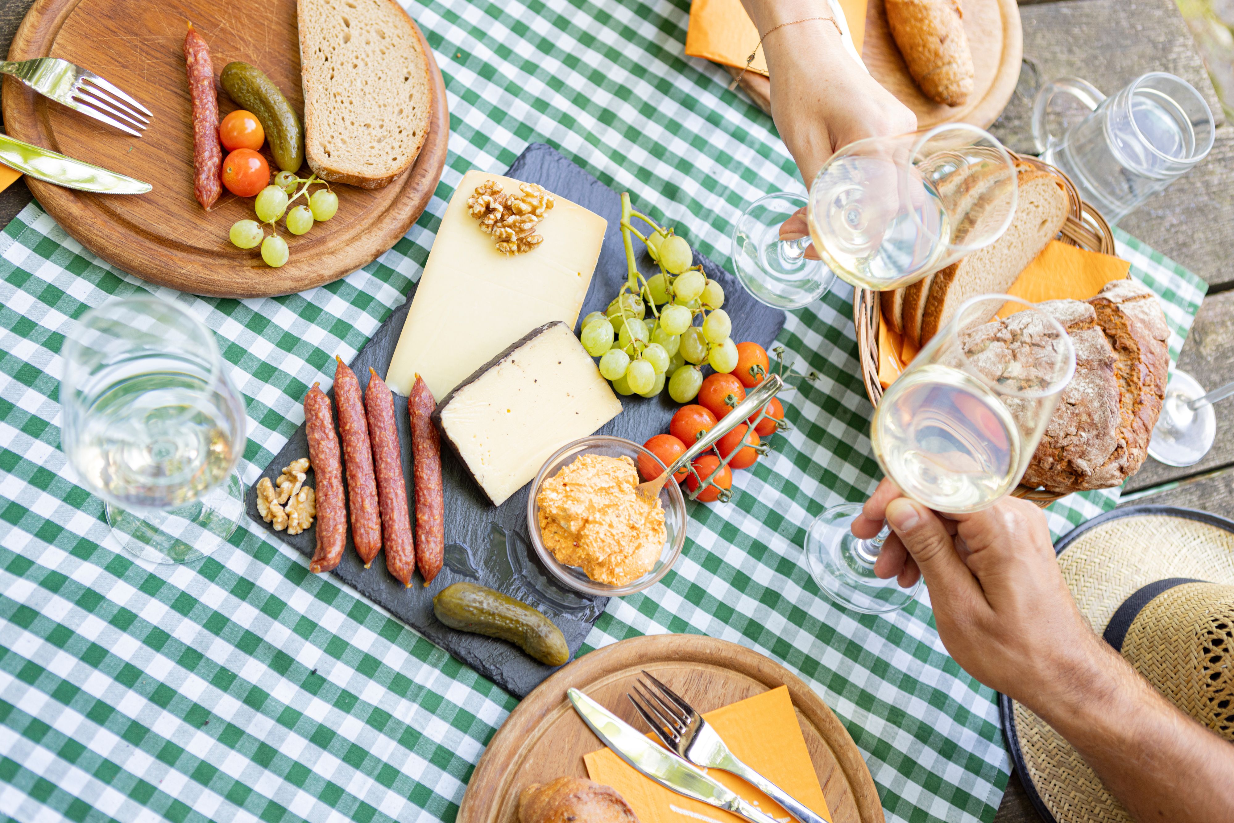 Picknicktisch mit Käse, Wurst, Brot und Wein auf kariertem Tuch.