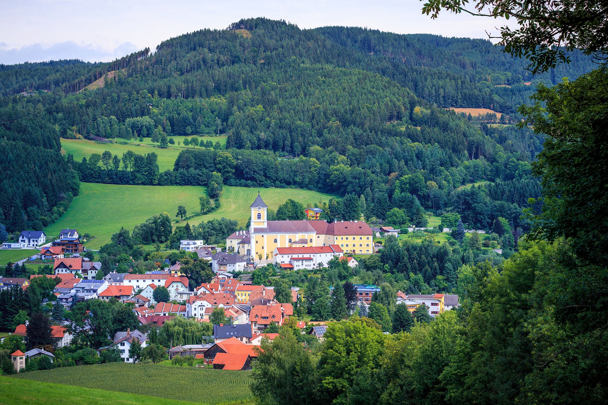 Landschaft mit Kirche und Kloster in einem Dorf, umgeben von Wäldern und Hügeln.