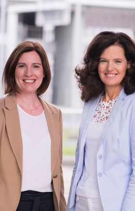 Edith Mader und Maria-Luise Fröch stehen lächelnd nebeneinander vor einem modernen Gebäude.