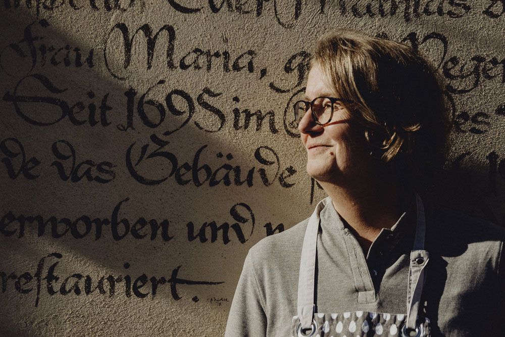 Person vor historischer Wand mit alter Schrift, lächelt im Sonnenlicht.
