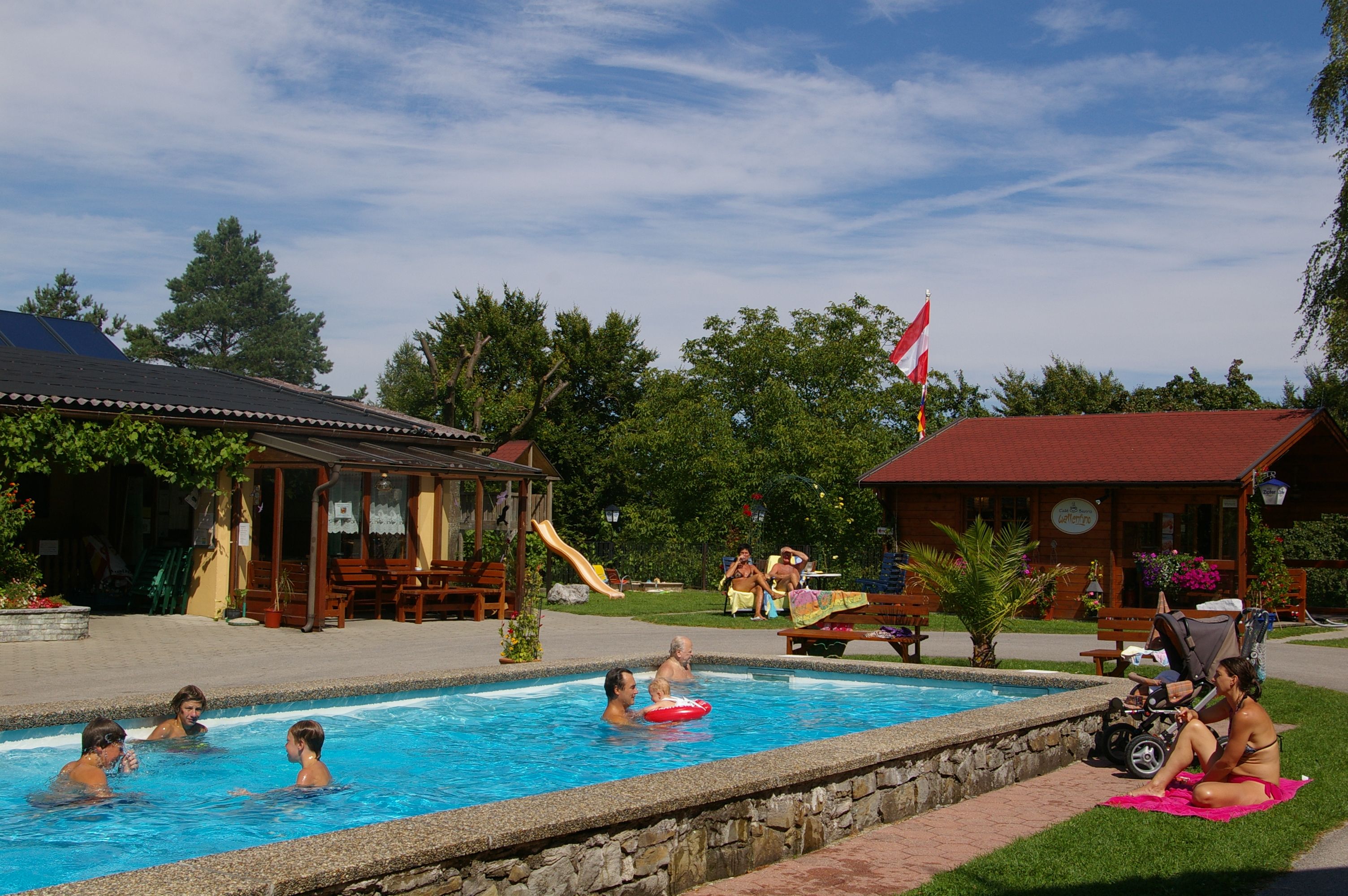 Menschen entspannen an einem kleinen Pool in einem Garten mit Holzhäusern und einer Rutsche.