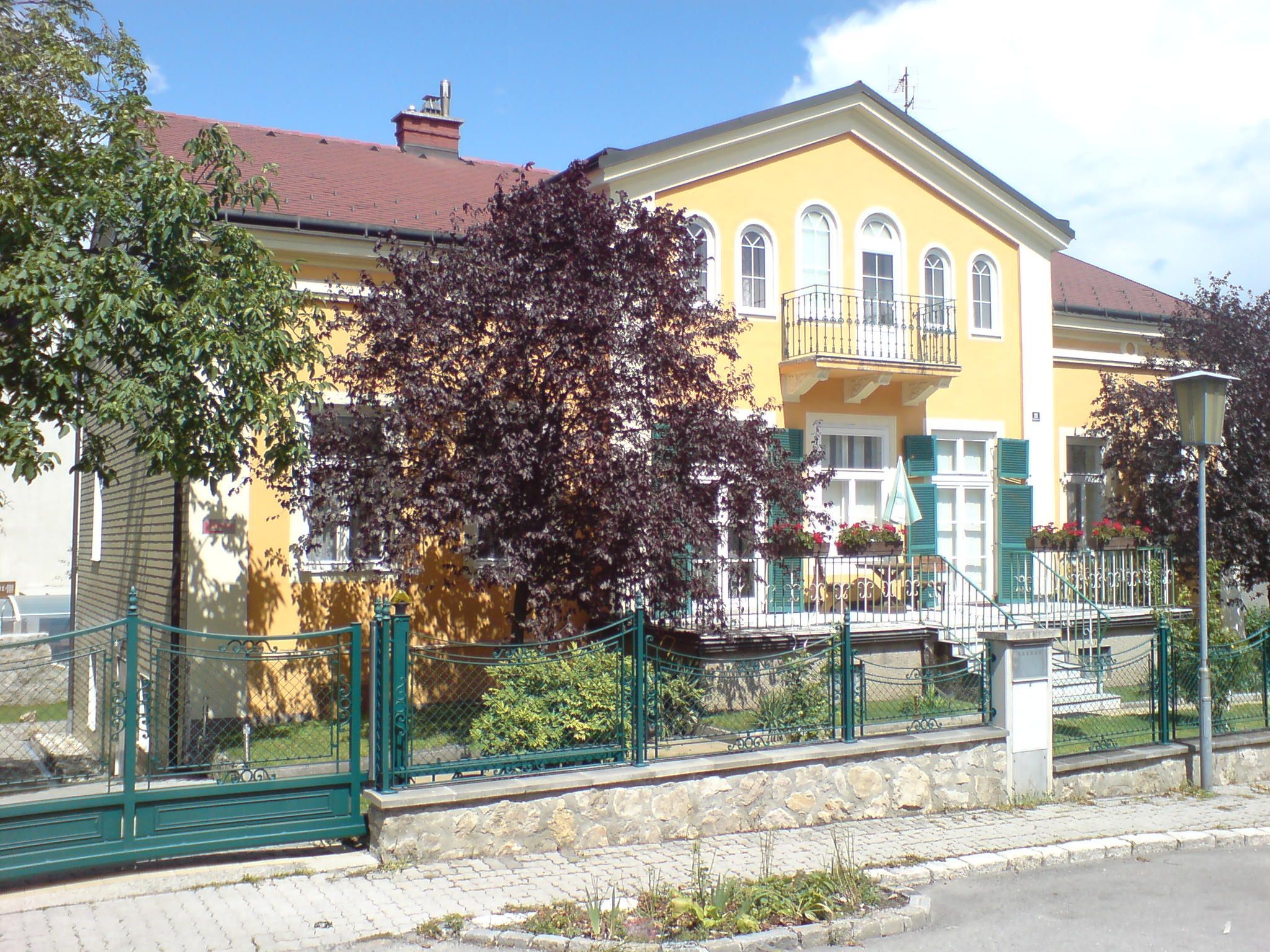 Gelbes Haus mit Balkon und grünem Zaun, umgeben von Bäumen.