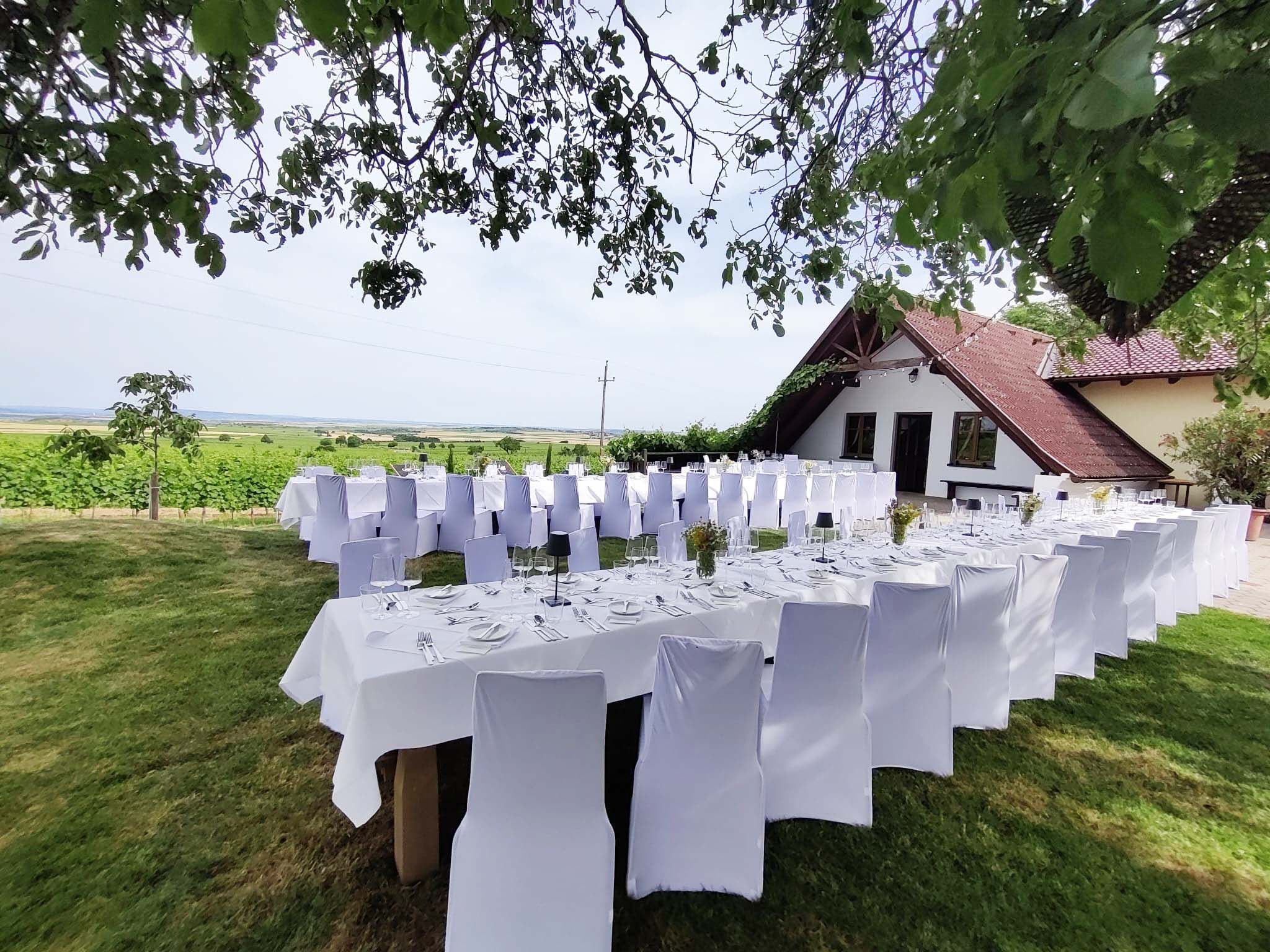 Gedeckte Tische im Freien mit weißen Stuhlhussen, umgeben von grüner Landschaft und einem Gebäude mit rotem Dach.