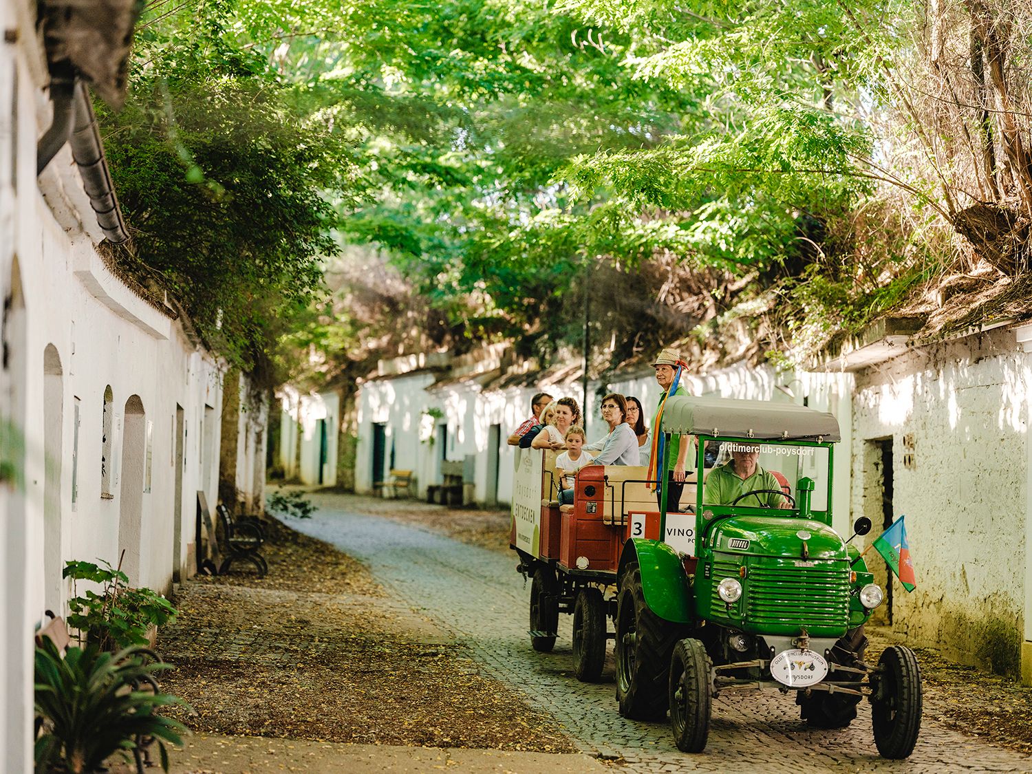 Ein grüner Oldtimer Traktor fährt durch eine Kellergasse, am Anhänger eine Gruppe Menschen