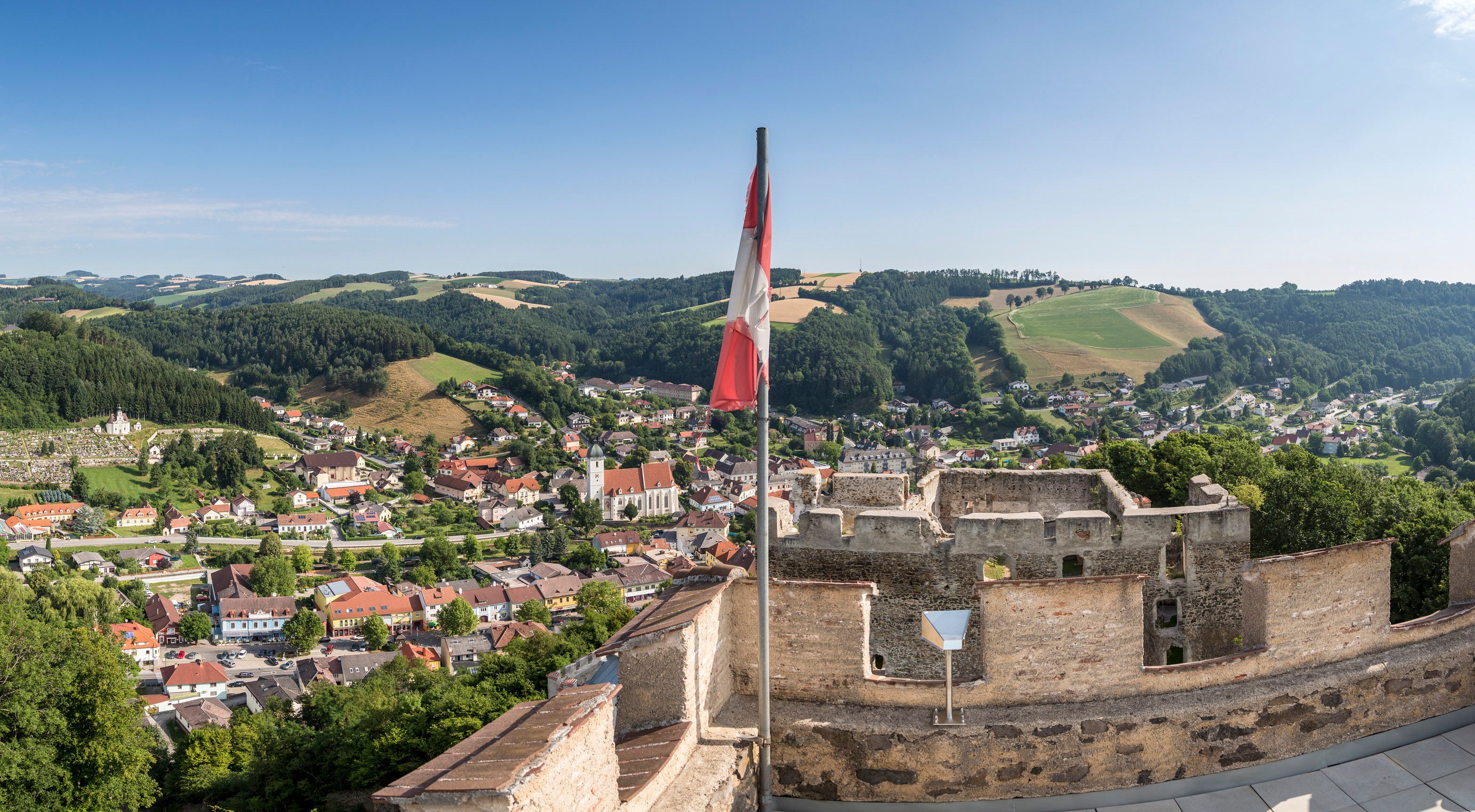 Panoramablick von einer Burgruine auf eine Stadt mit Hügeln im Hintergrund und einer österreichischen Flagge im Vordergrund.