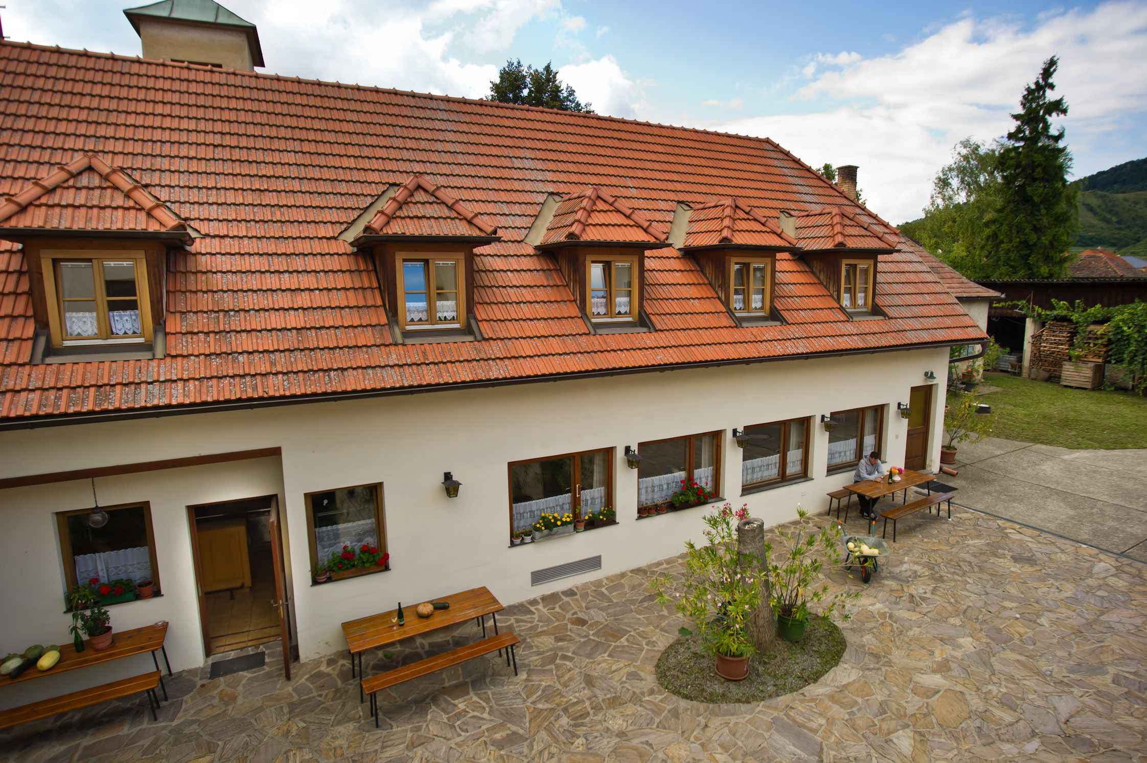 Ein traditionelles Gebäude mit rotem Ziegeldach und Dachfenstern, umgeben von einem gepflasterten Innenhof mit Tischen und Pflanzen.