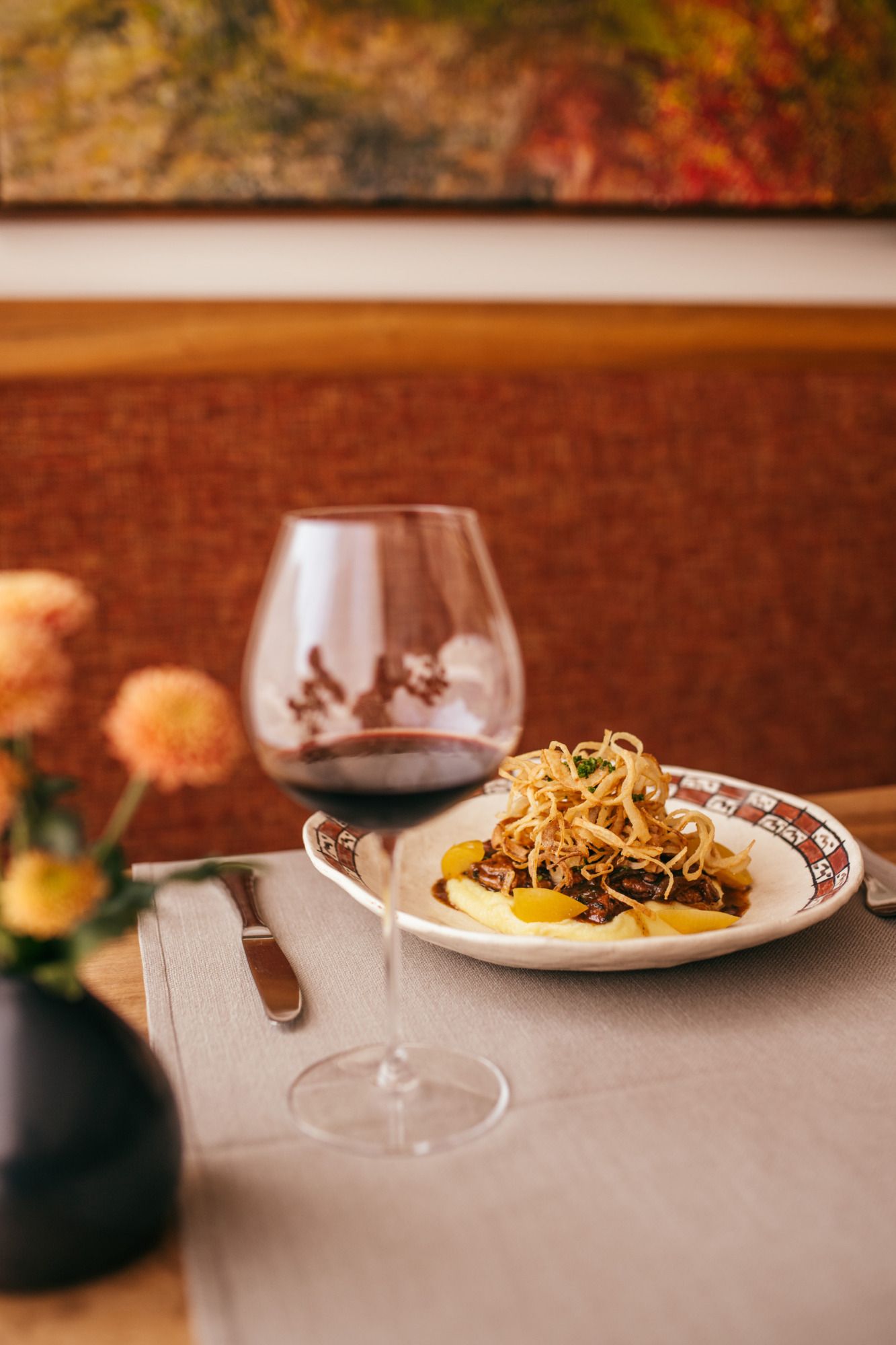 Ein Teller mit glasierter Kalbsleber auf Kartoffelpüree, garniert mit Röstzwiebeln, neben einem Glas Rotwein auf einem Tisch.