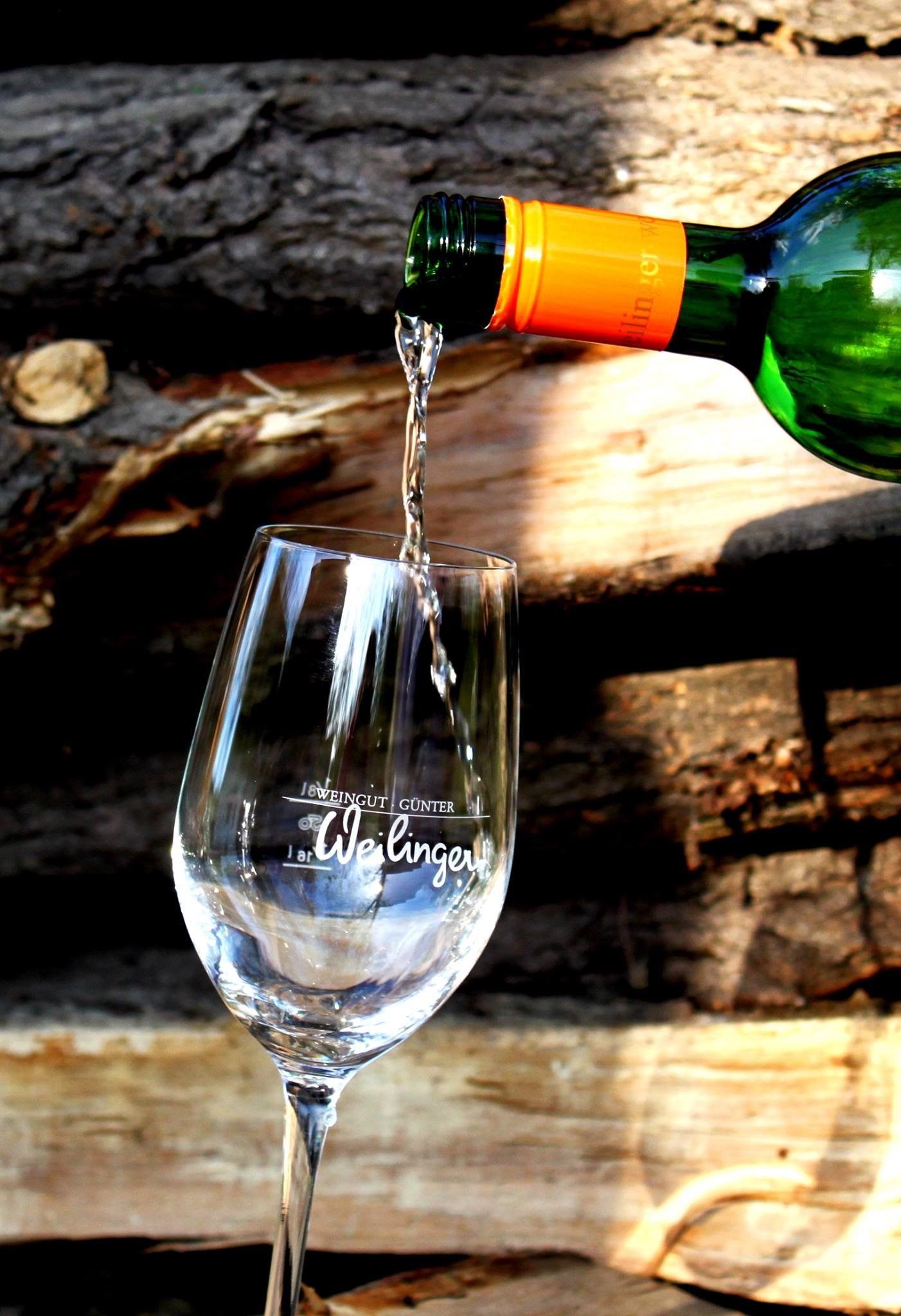 Ein Glas Wein wird aus einer Flasche mit orangefarbenem Verschluss eingeschenkt. Im Hintergrund sind Holzscheite zu sehen.