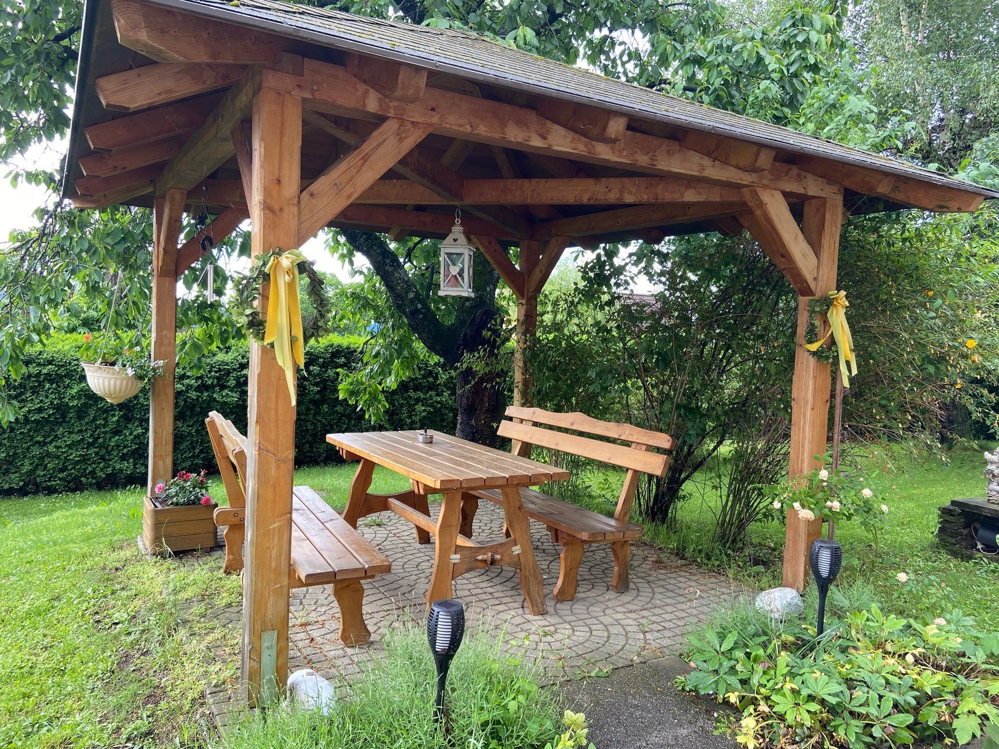 Holzlaube im Garten mit Tisch und Bänken, umgeben von Pflanzen.