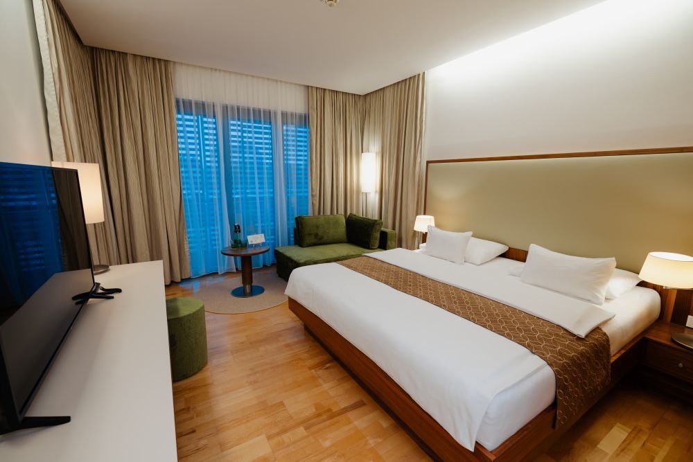 Modernes Hotelzimmer mit großem Bett, Sofa, Fernseher und Vorhängen vor einem Fenster.