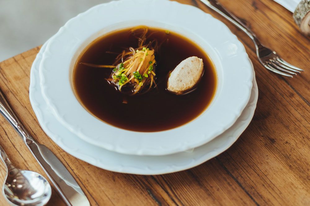 Eine klare Rindssuppe mit Gemüse und Knödel in einem weißen Teller auf einem Holztisch.