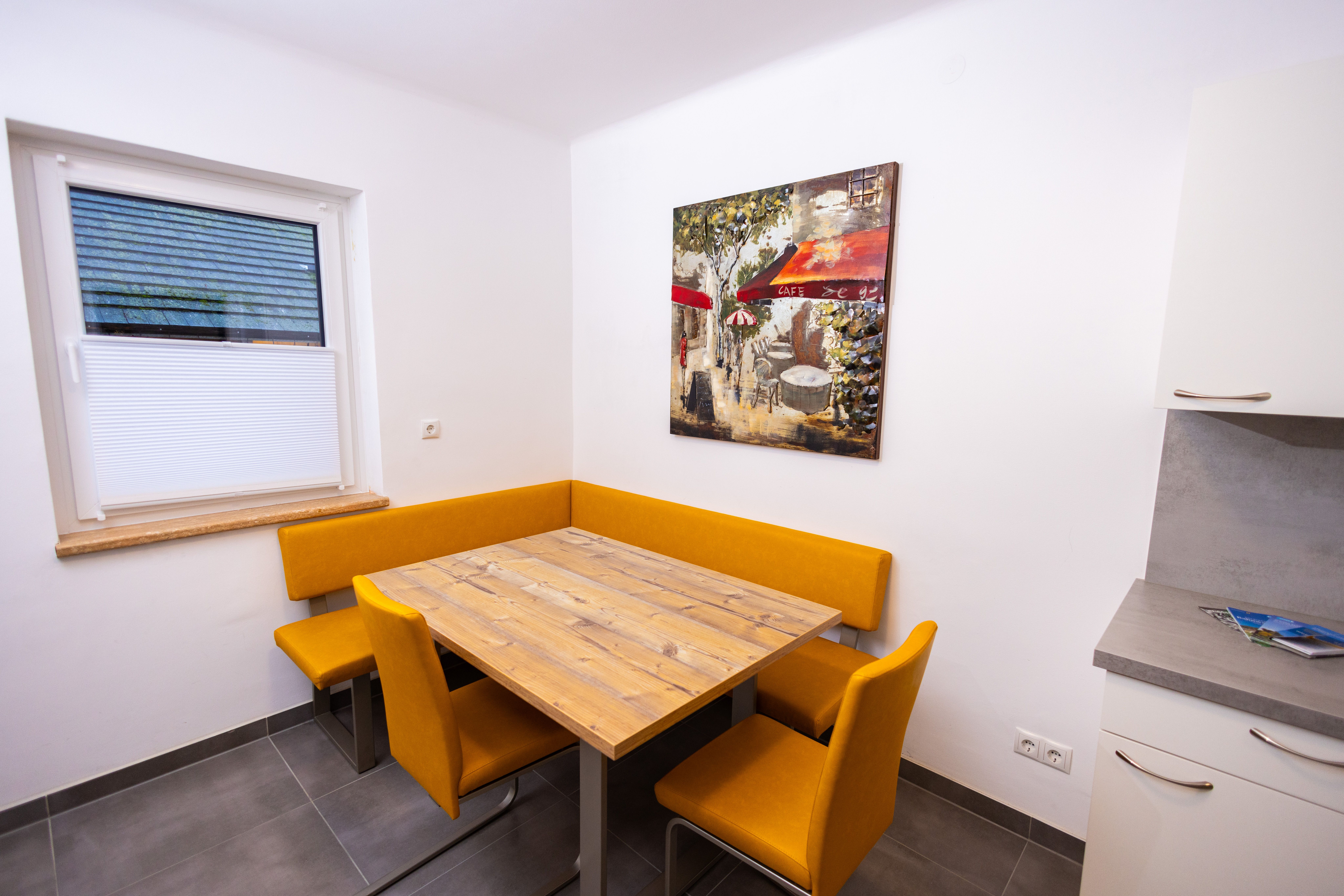 Essbereich mit Holztisch, gelben Stühlen und Wandbild eines Cafés.