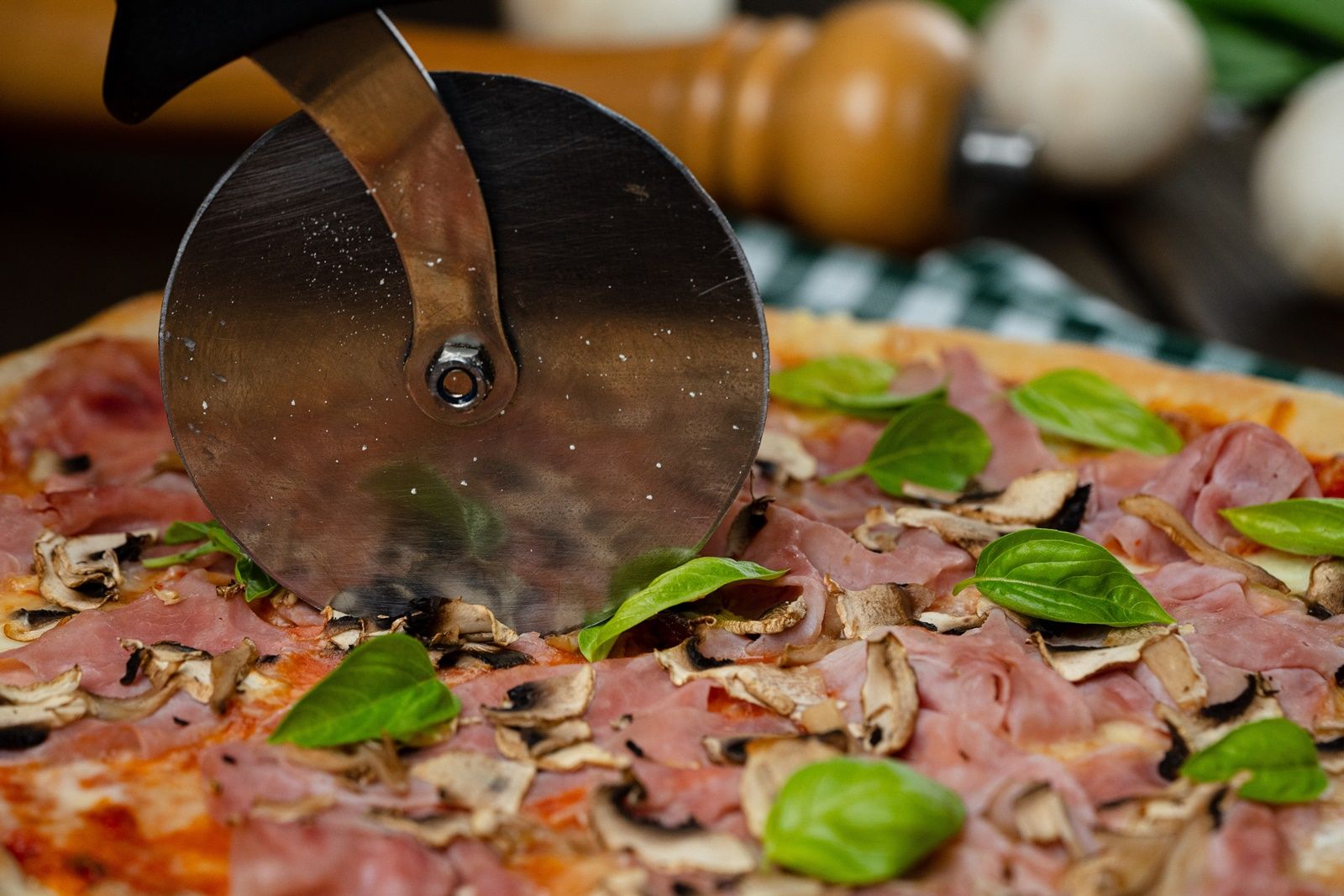 Nahaufnahme einer Pizza mit Schinken, Pilzen und Basilikum, die mit einem Pizzaschneider geschnitten wird.