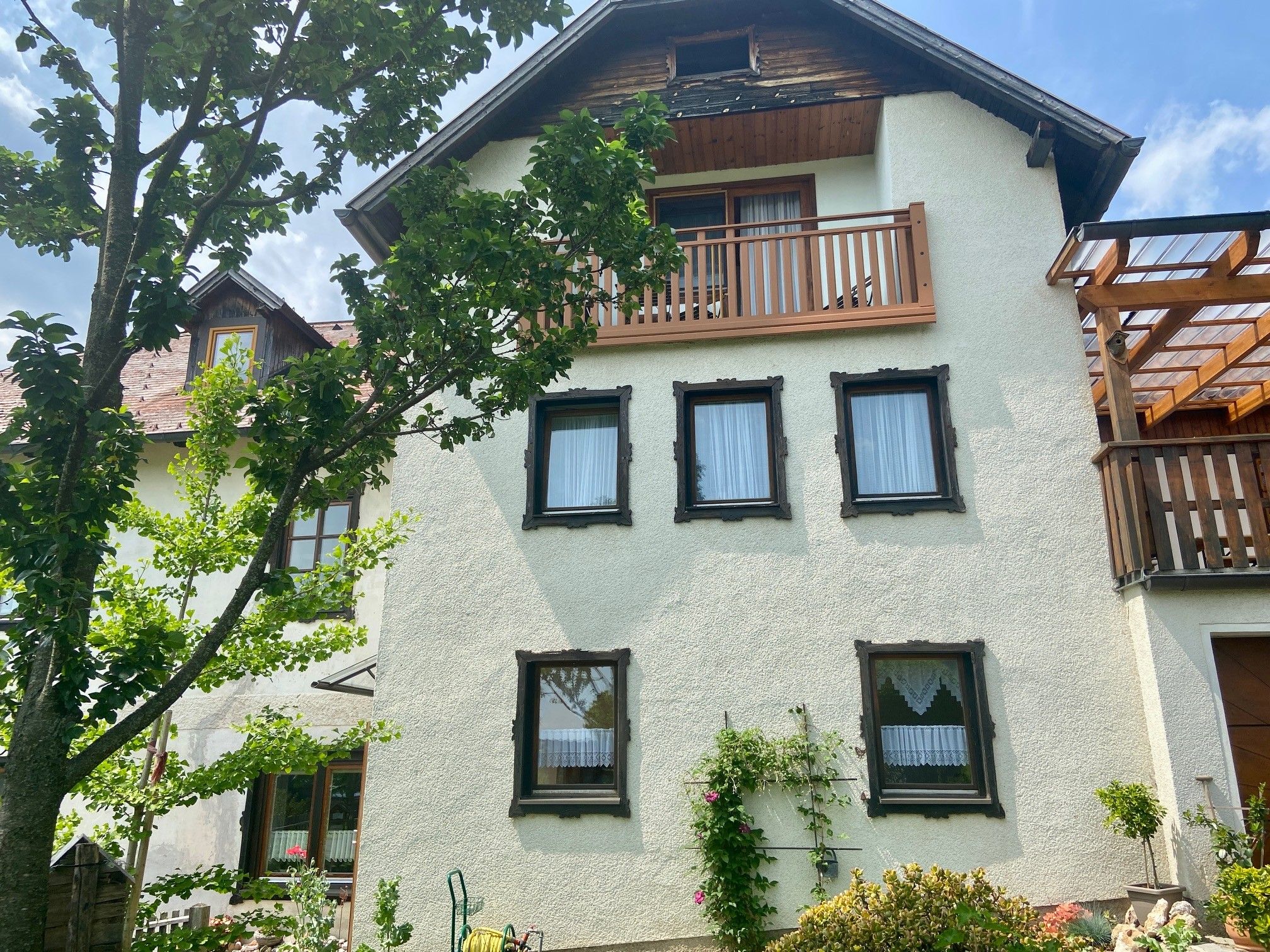 Ein traditionelles Haus mit weißen Wänden, Holzverkleidung und Balkon, umgeben von grünen Bäumen und Pflanzen.