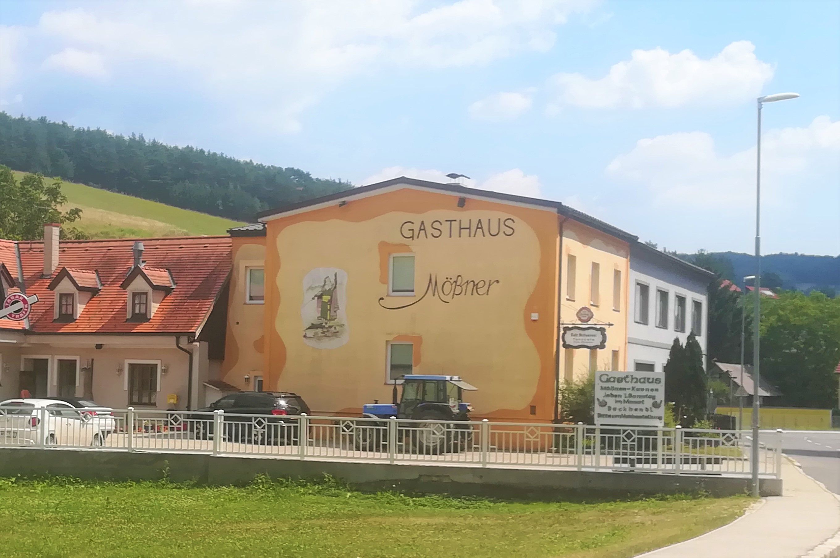 Außenansicht des Gasthauses Mößner, gelb-orange gestrichen, in einer ländlichen Umgebung mit blauer Traktor und Autos vor dem Gebäude.