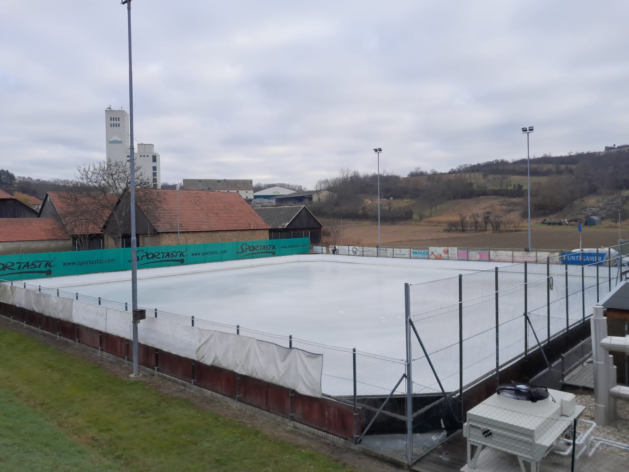 Ein leerer Eislaufplatz mit umliegenden Gebäuden und Hügeln im Hintergrund.