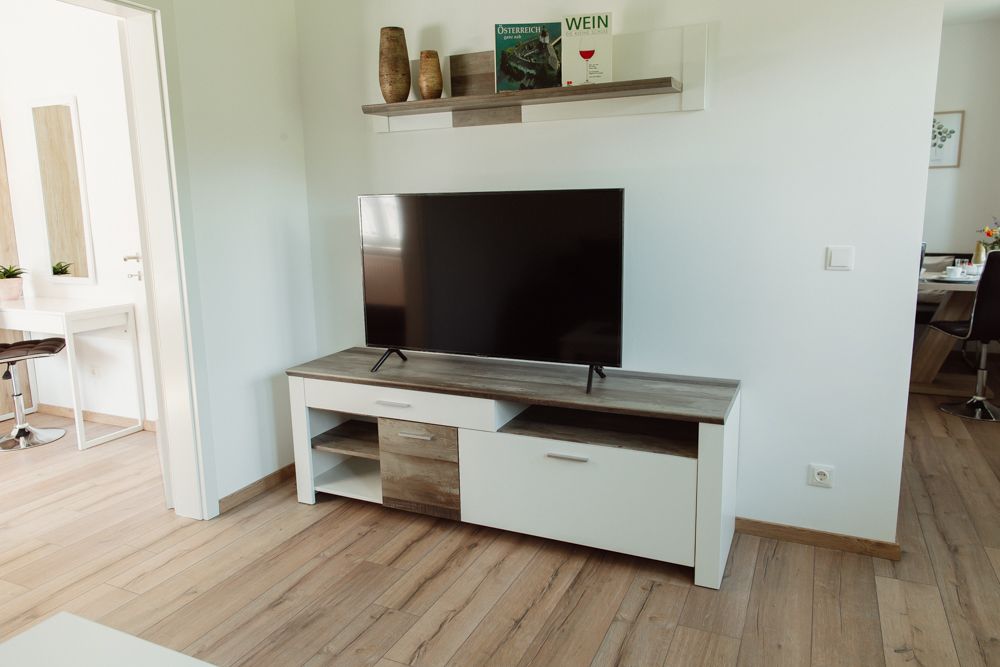 Wohnzimmer mit Fernseher auf einem weißen TV-Schrank, darüber ein Regal mit Dekorationen.