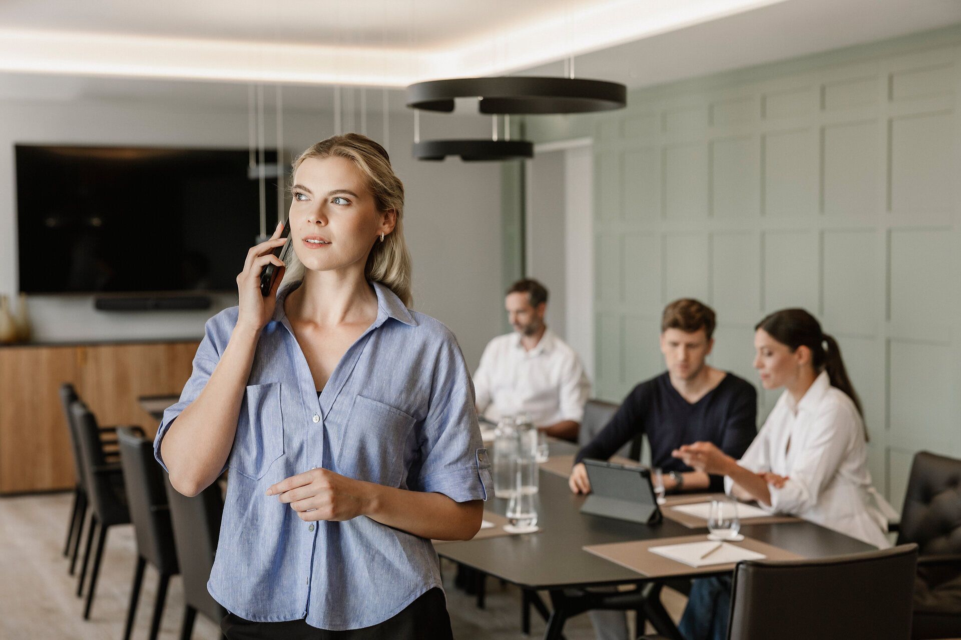 Eine Frau im Vordergrund am Telefon, drei Personen in einem Business Meeting in modernem Setting an einem schwarzen Konferenztisch im Mörwald Gutshof.