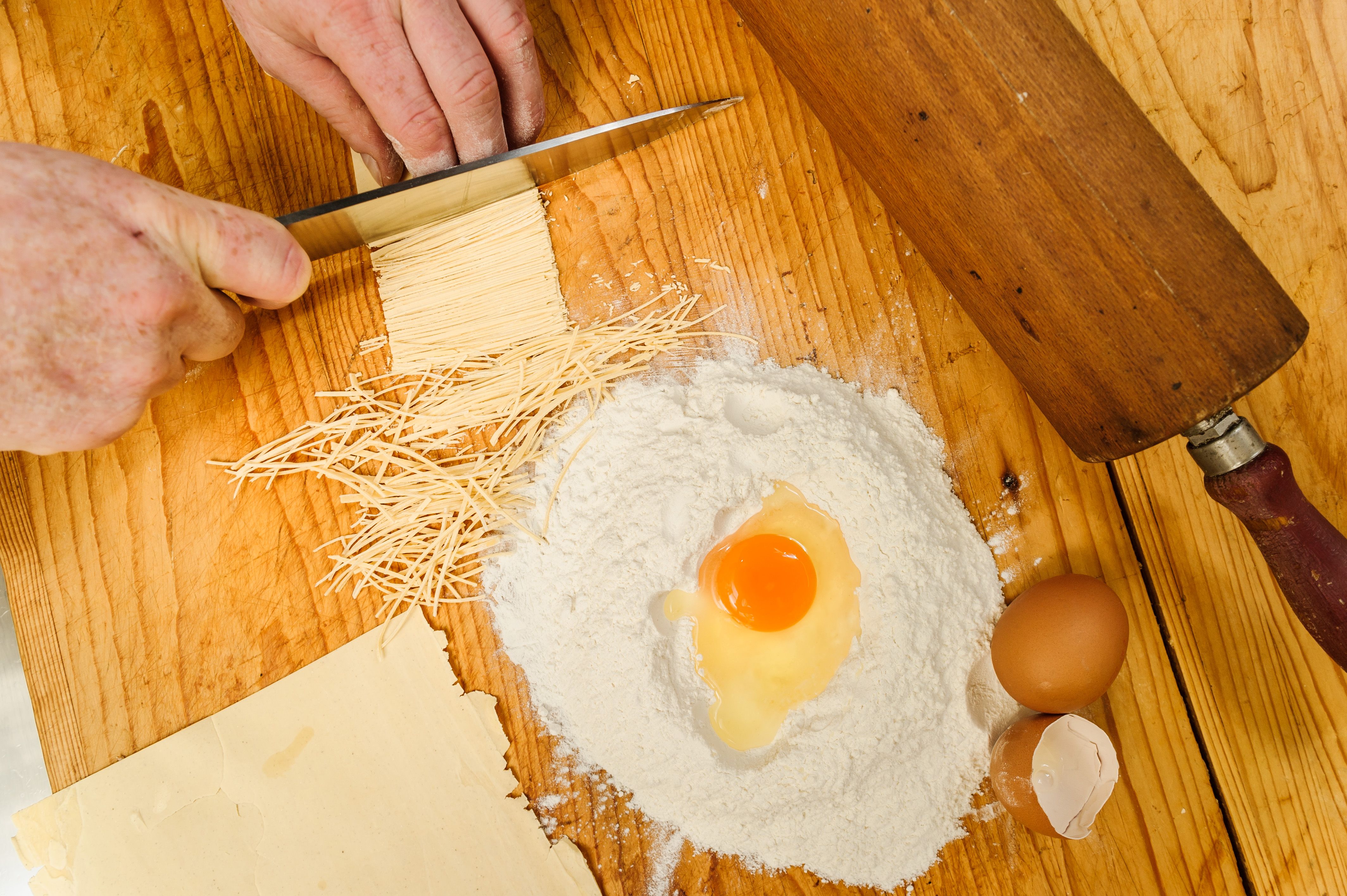 Nahaufnahme von Händen, die Nudelteig schneiden, mit Mehl, Ei und Nudelholz auf einem Holztisch.