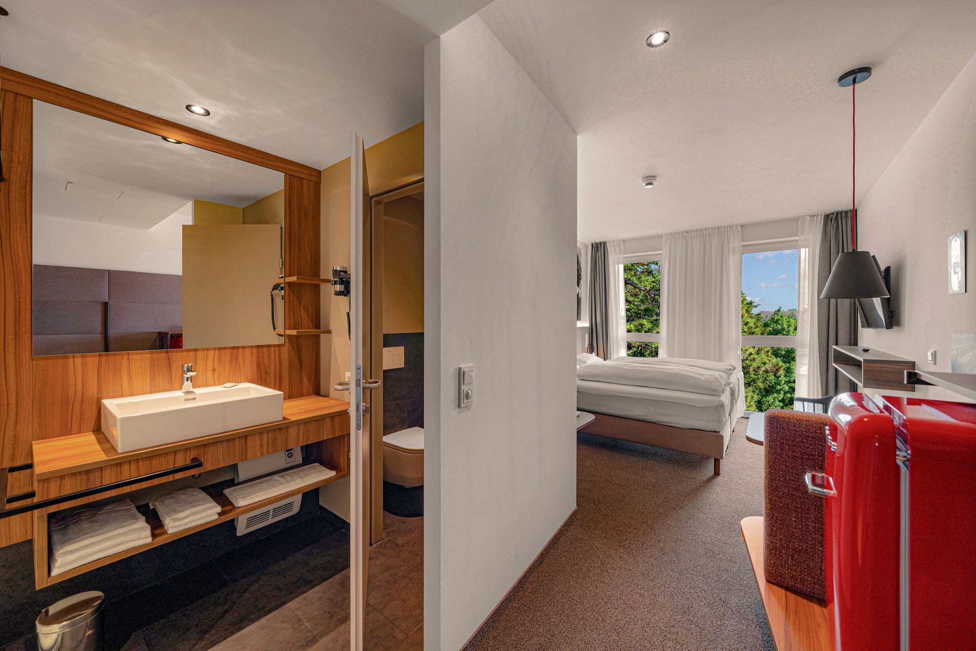 Innenansicht eines modernen Hotelzimmers mit Bad, Bett und rotem Kühlschrank.