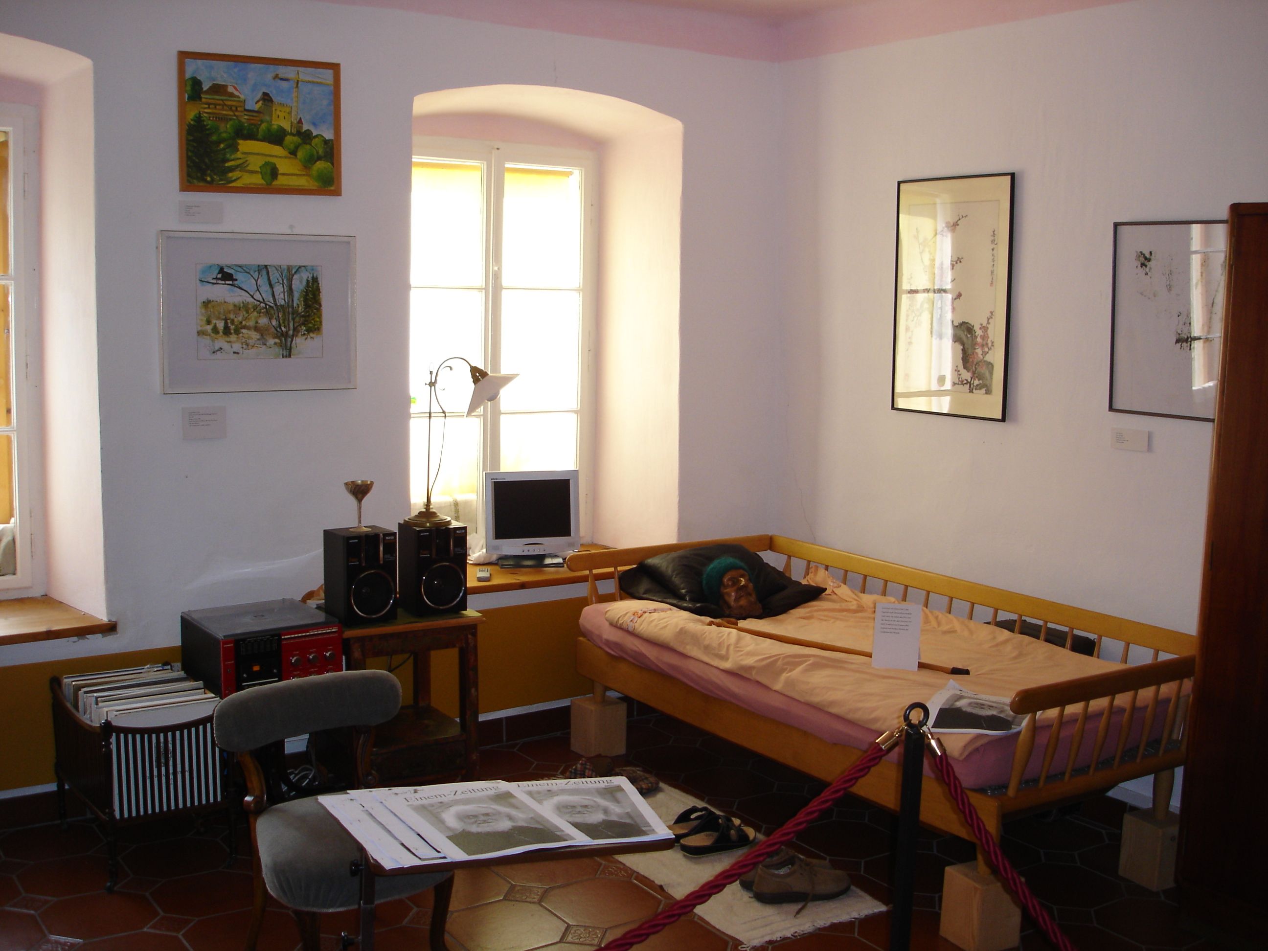 Innenansicht eines Museumszimmers mit Bett, Bildern an der Wand und einem kleinen Schreibtisch mit Computer.