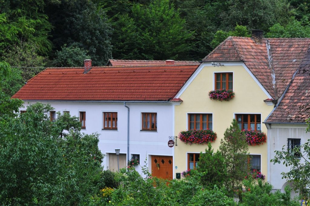 Ein traditionelles Haus mit roten Dachziegeln und blühenden Blumenkästen vor den Fenstern, umgeben von grüner Vegetation.