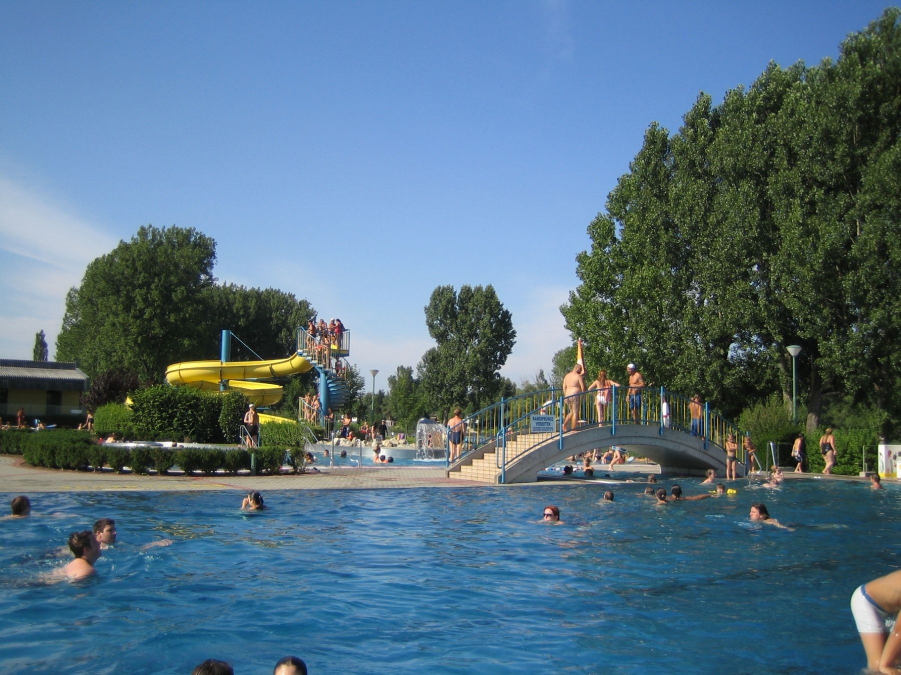 Freibad mit Wasserrutsche und Brücke, viele Menschen im Wasser.
