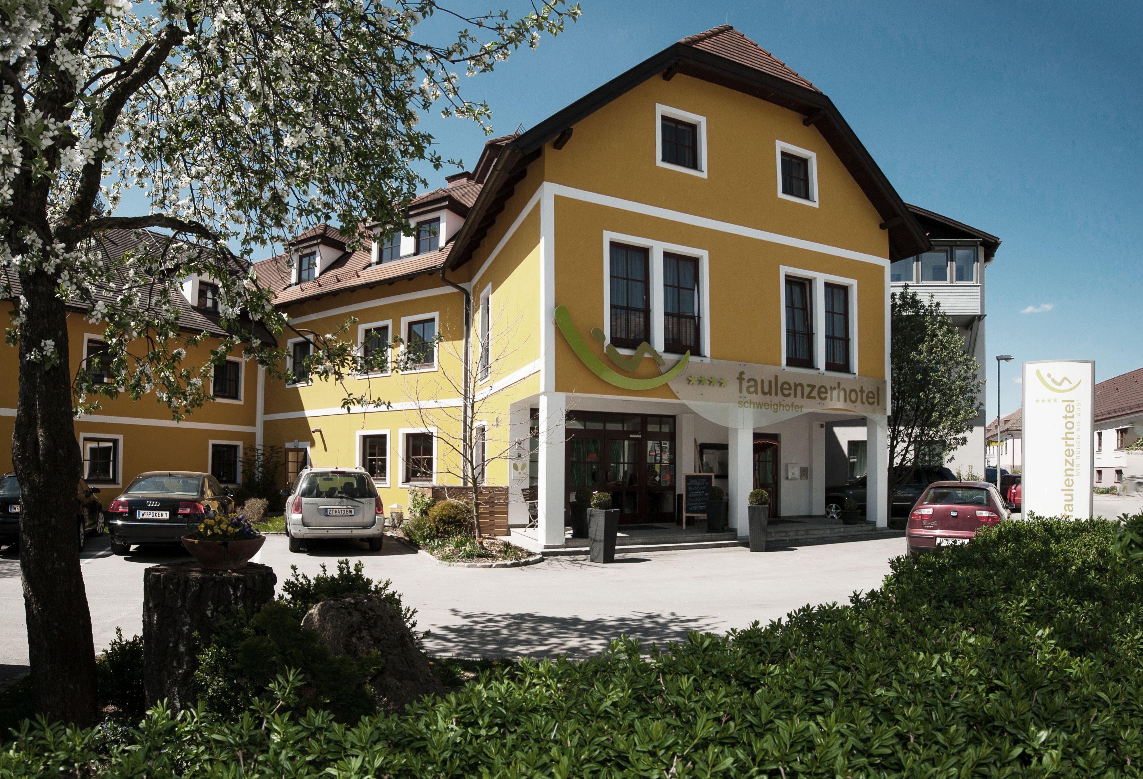Gelbes Hotelgebäude mit Autos im Vordergrund und blühendem Baum.