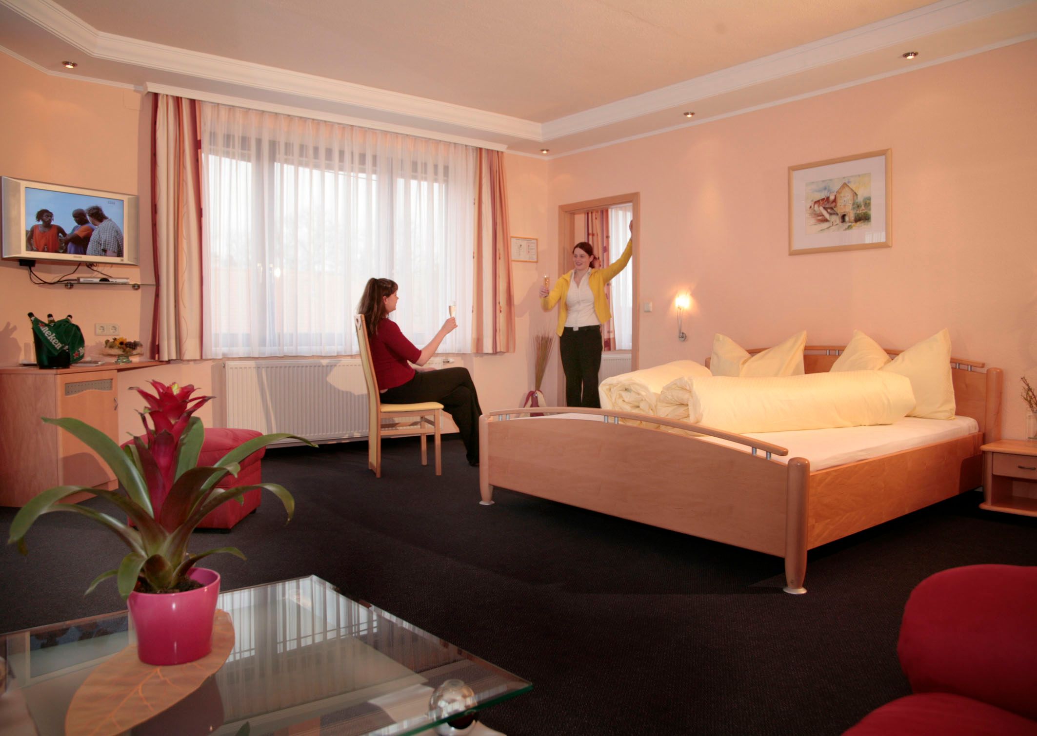 Zwei Frauen in einem Hotelzimmer mit Doppelbett, eine sitzt, die andere steht mit erhobenem Glas.