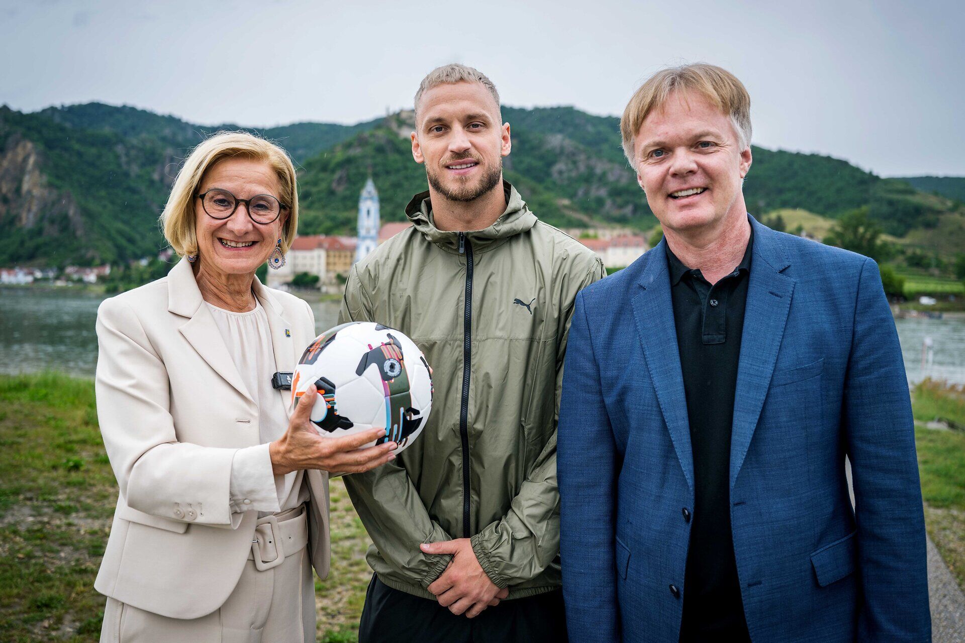 Landeshauptfrau Johanna Mikl-Leitner hält einen Fußball in den Händen. Neben ihr stehen Fußballprofi und Niederösterreich-Testimonial Marko Arnautović sowie Michael Duscher, Geschäftsführer der Niederösterreich Werbung. Im Hintergrund ist die Wachauer Landschaft zu sehen.
