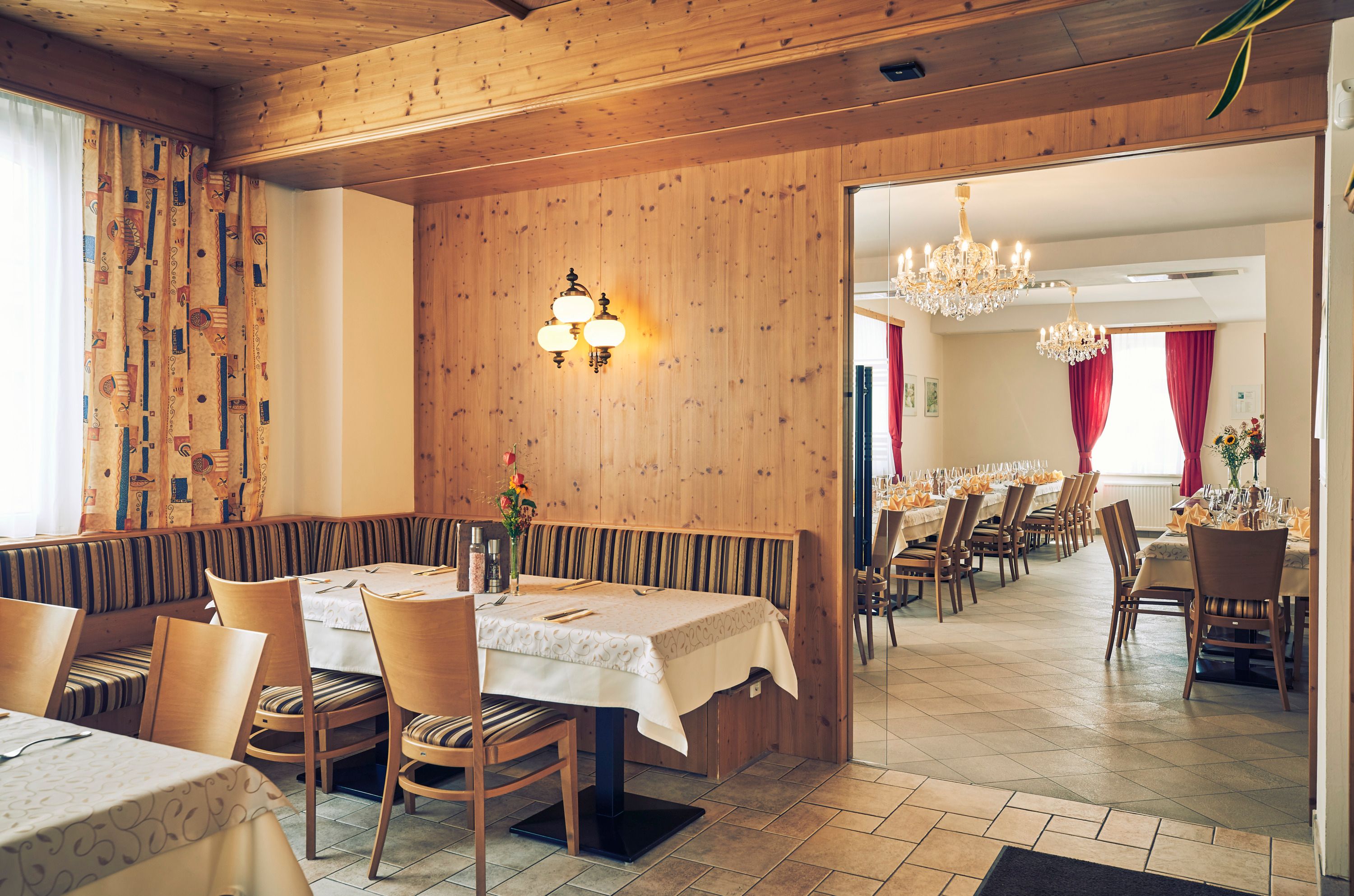 Ein gemütliches Gastzimmer mit Holzwänden und ein angrenzender Festsaal mit Kronleuchtern und gedeckten Tischen.