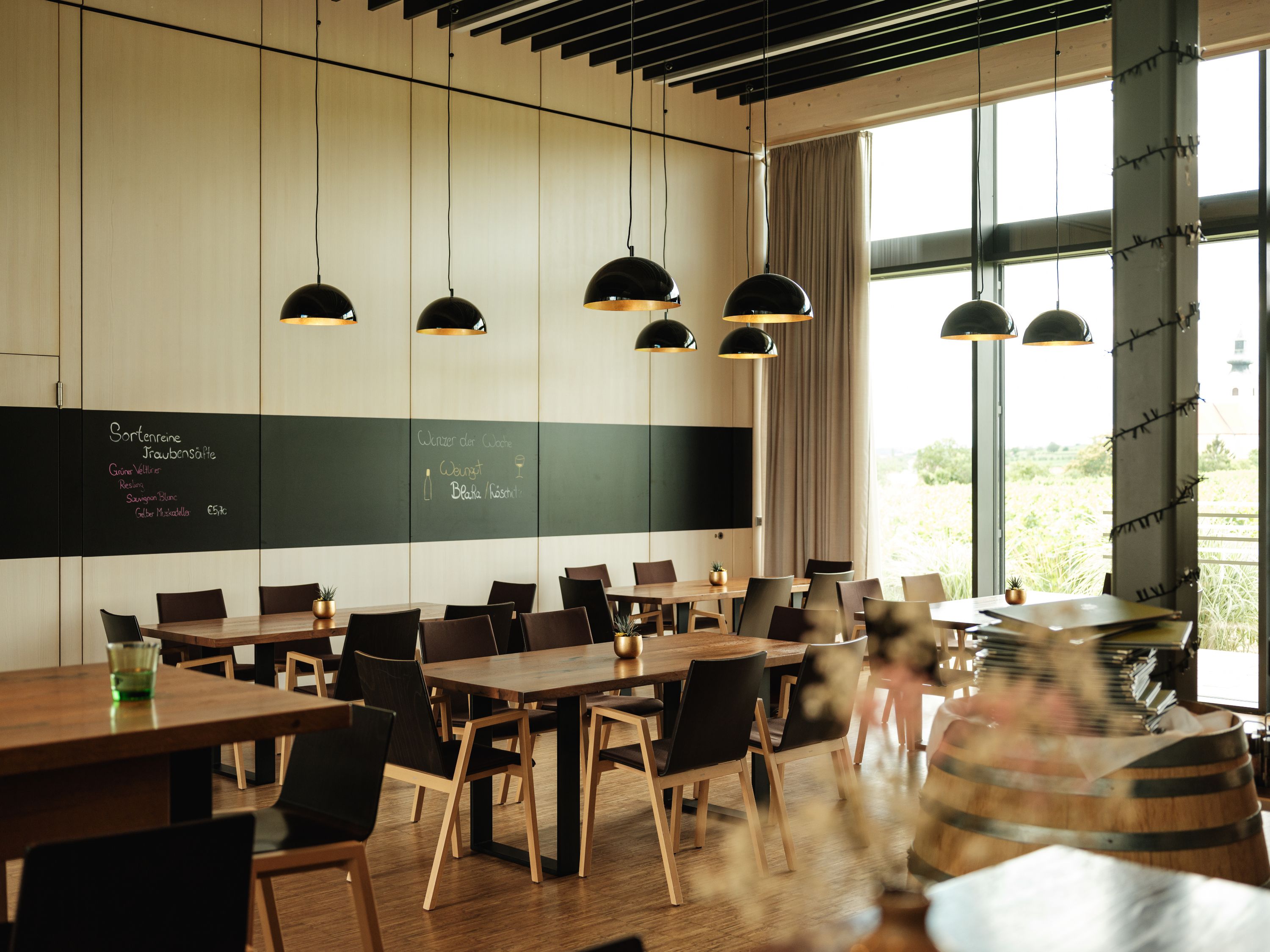 Modernes Restaurant mit Holztischen, schwarzen Stühlen und großen Fenstern. Schwarze Hängelampen und eine Tafel an der Wand.