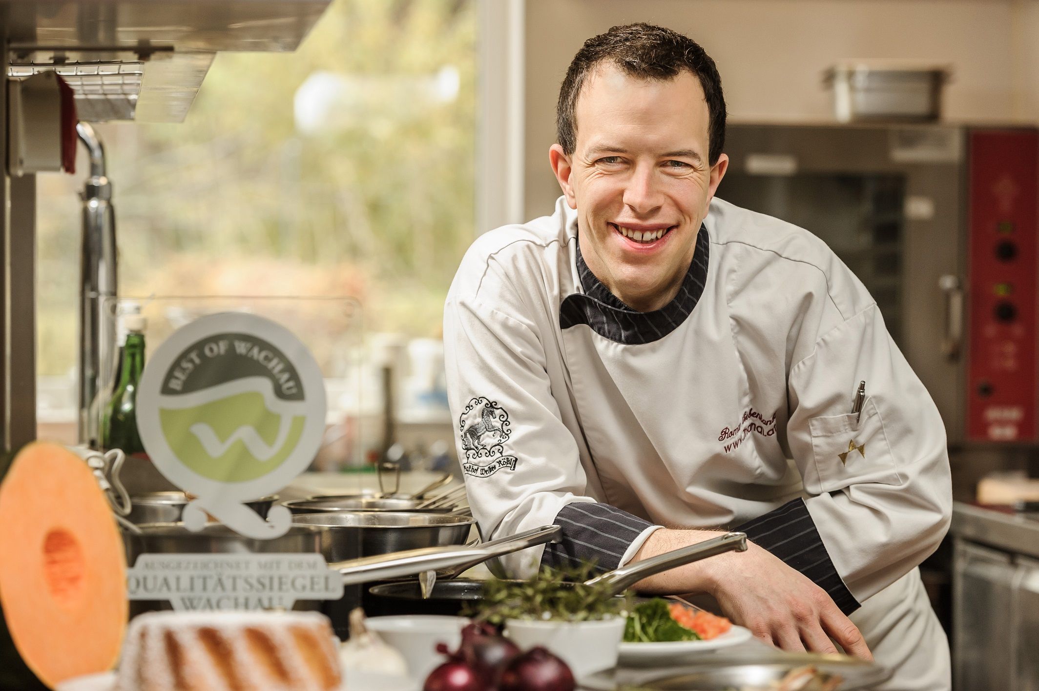 Ein Koch in weißer Uniform lächelt in einer Küche, umgeben von frischen Zutaten und einem Qualitätssiegel der Wachau.