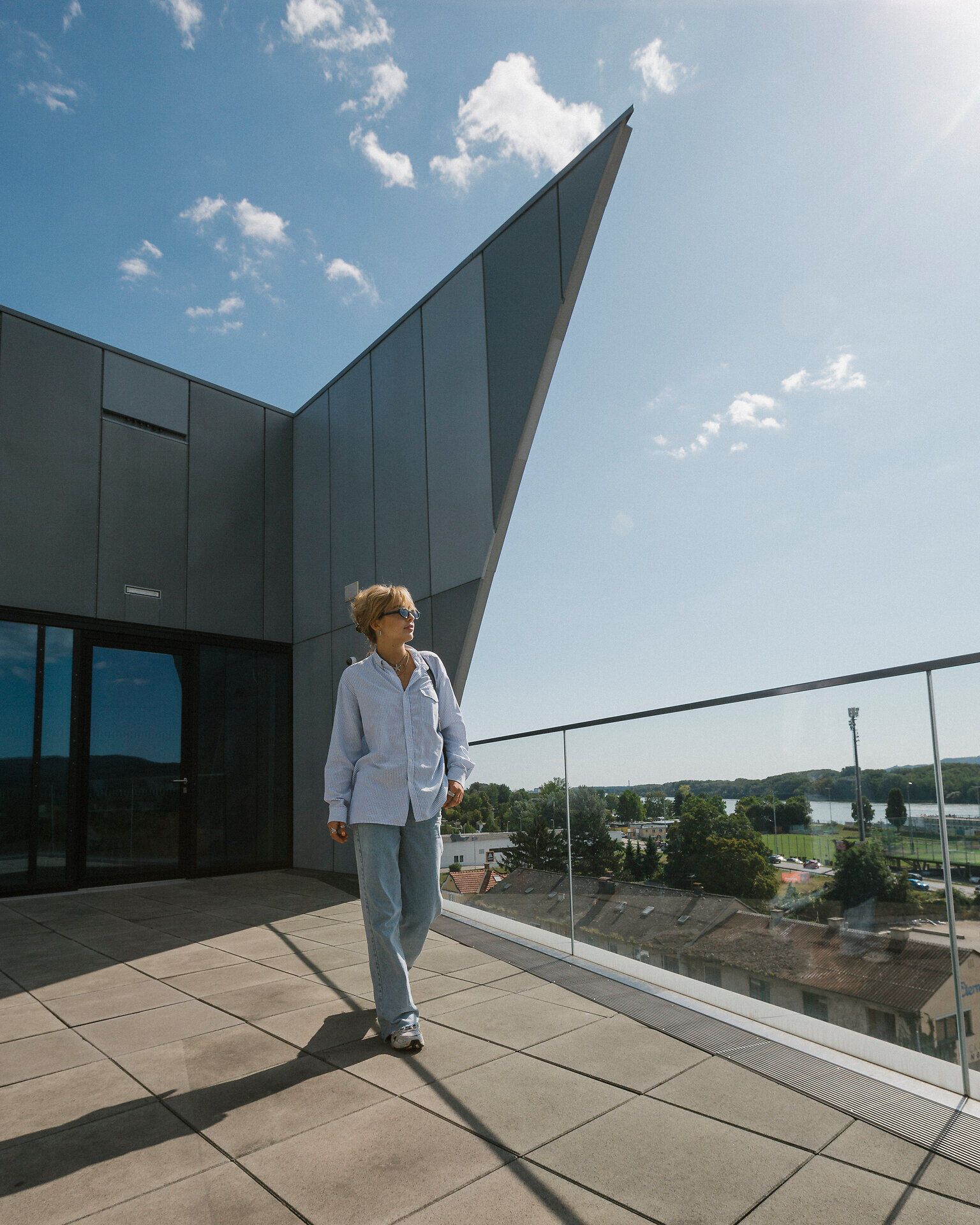 Person in hellem Hemd und Jeans geht über eine Dachterrasse mit Glasgeländer; im Hintergrund ein modernes Gebäude mit markanter, spitz zulaufender Dachkante und blauer Himmel mit kleinen Wolken.