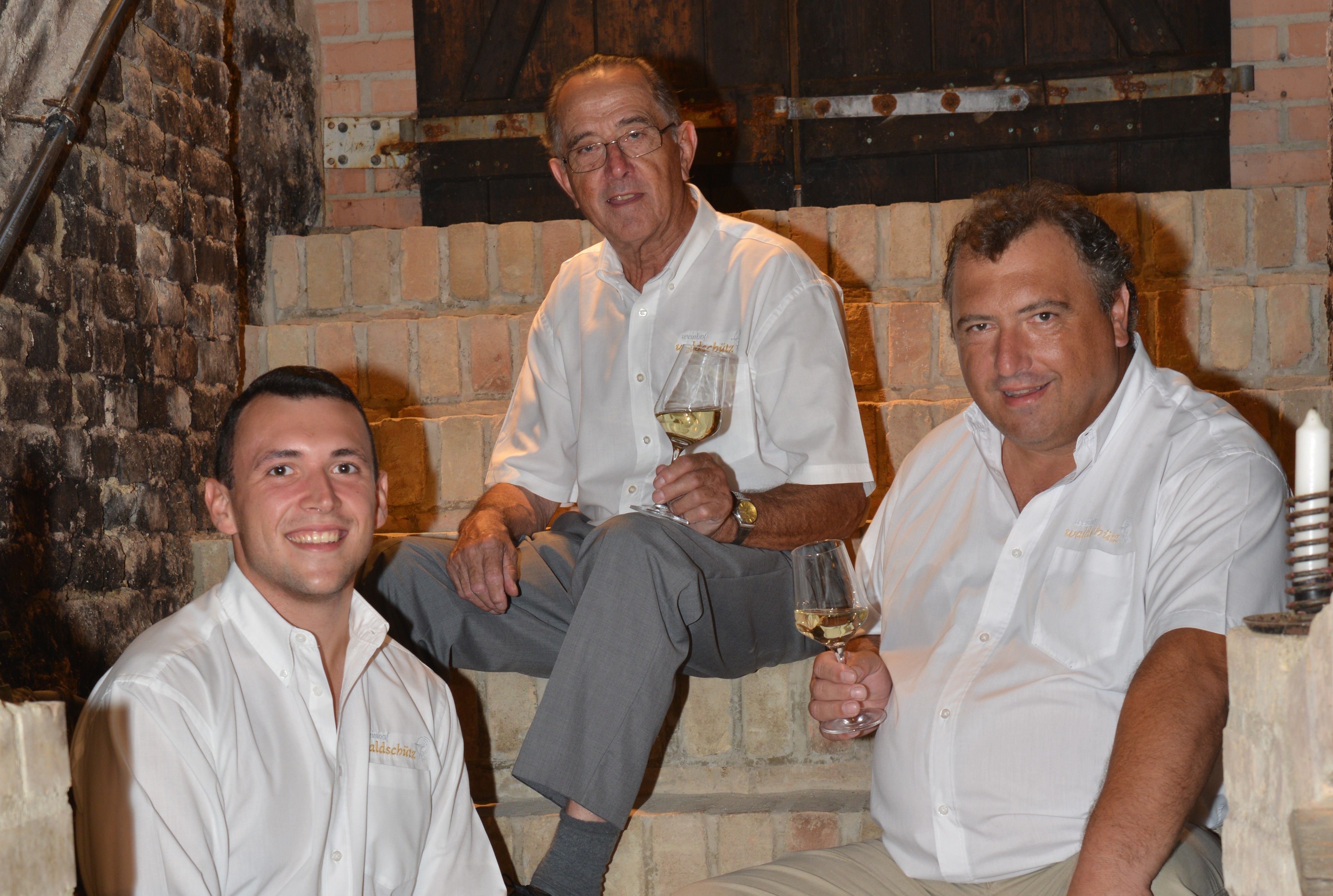 Drei Männer in weißen Hemden sitzen auf einer Steintreppe und halten Weingläser.