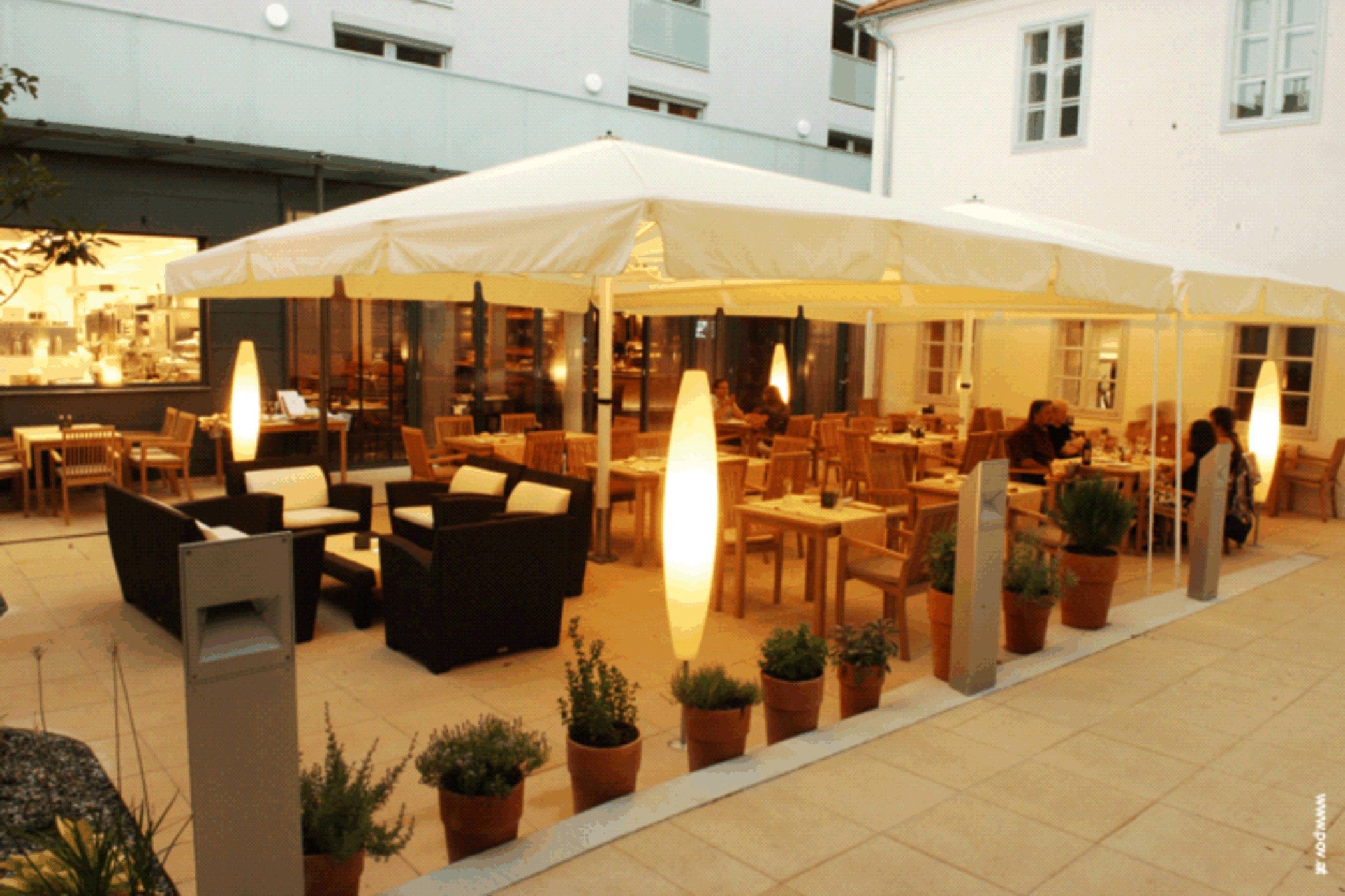 Ein beleuchteter Gastgarten mit Tischen, Stühlen und Pflanzen in Töpfen, umgeben von Gebäuden.