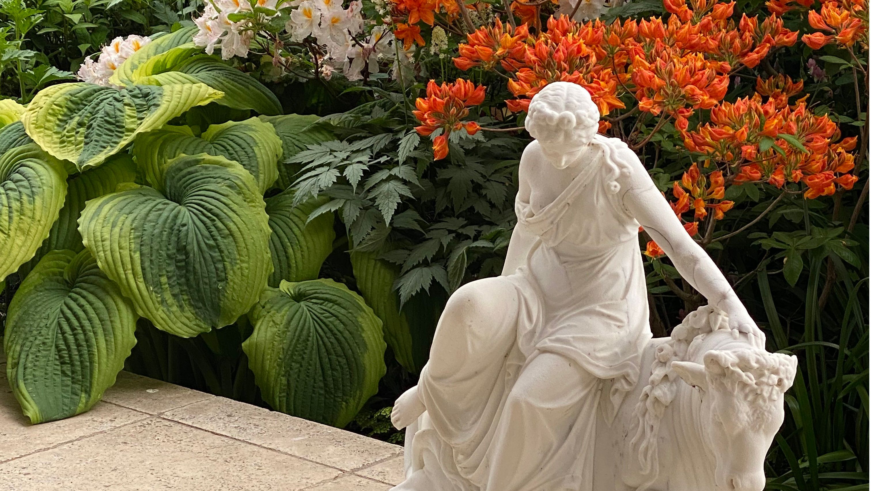 Weiße Statue einer Frau neben großen grünen Blättern und orangefarbenen Blumen.