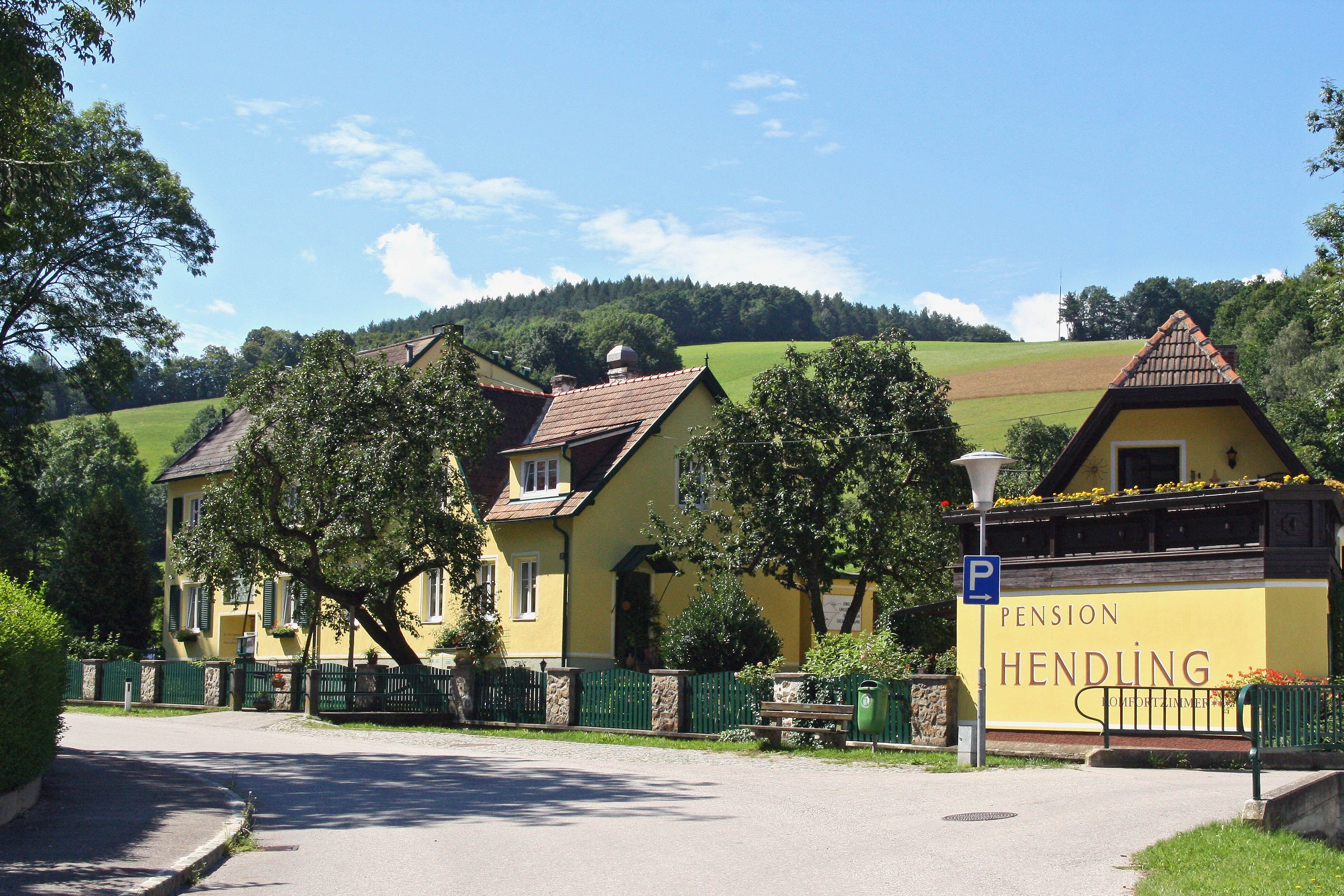 Gelbes Gebäude der Pension Hendling in ländlicher Umgebung mit grünen Hügeln im Hintergrund.