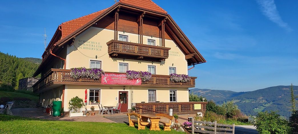 Ein traditionelles Gasthaus mit Holzbalkonen und Blumen, umgeben von grünen Hügeln und blauem Himmel.