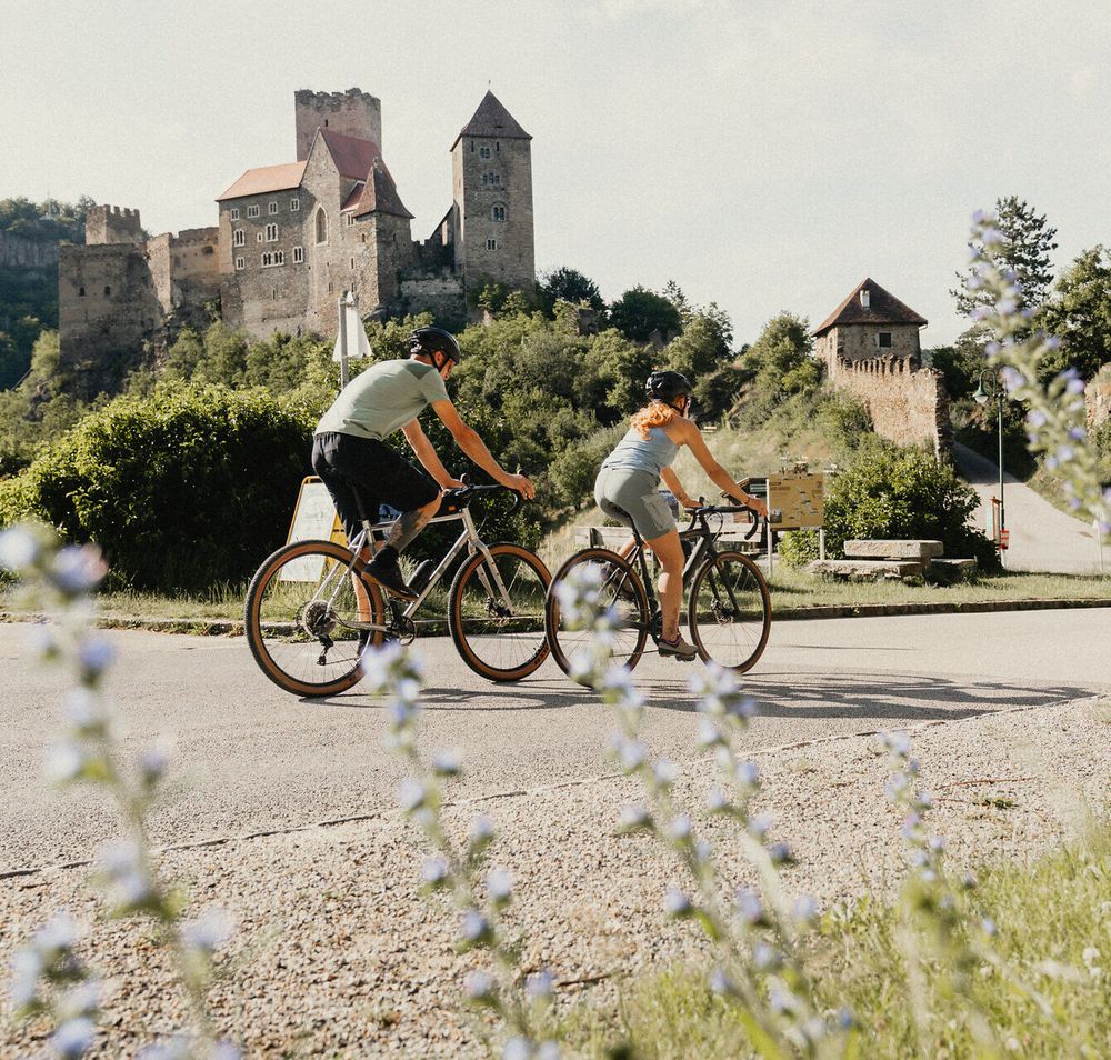 Zwei Radfahrer fahren entlang eine Straße, hinter ihnen die Burg Hardegg, im Vordergrund Blumen