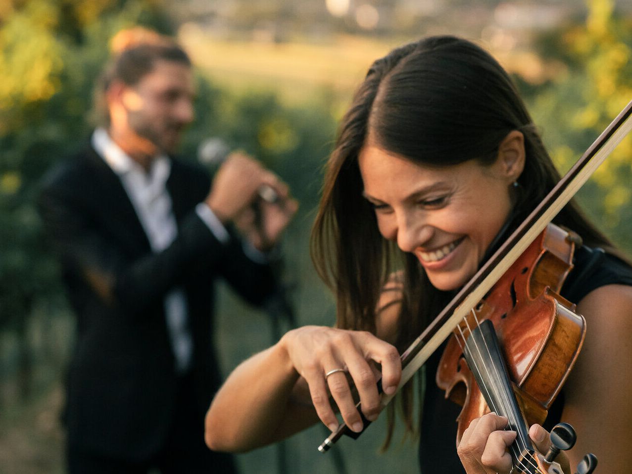 Eine Frau spielt draußen fröhlich Geige, während ein Mann im Hintergrund eins Mikrofon singt. Beide sind elegant gekleidet, und die Umgebung scheint ein weinbergähnliches Setting zu sein.