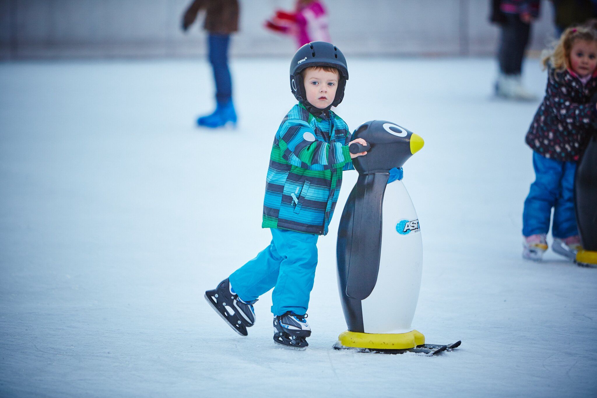 Ein Kind mit Helm und bunter Jacke hält sich an einer Pinguin-Eislaufhilfe auf einer Eisbahn fest.