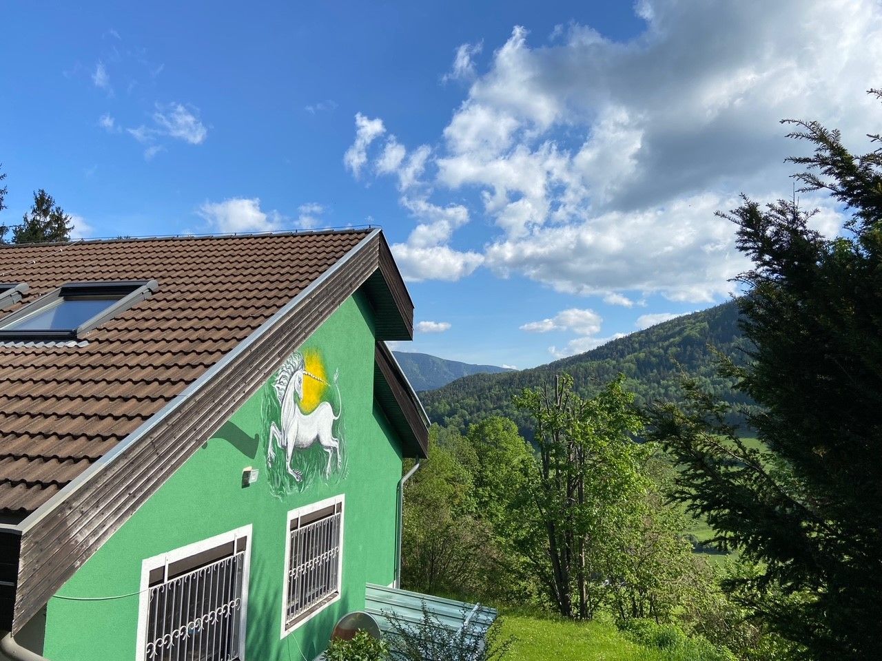 Grünes Haus mit Einhornbild an der Wand, umgeben von Bäumen und Bergen unter blauem Himmel.
