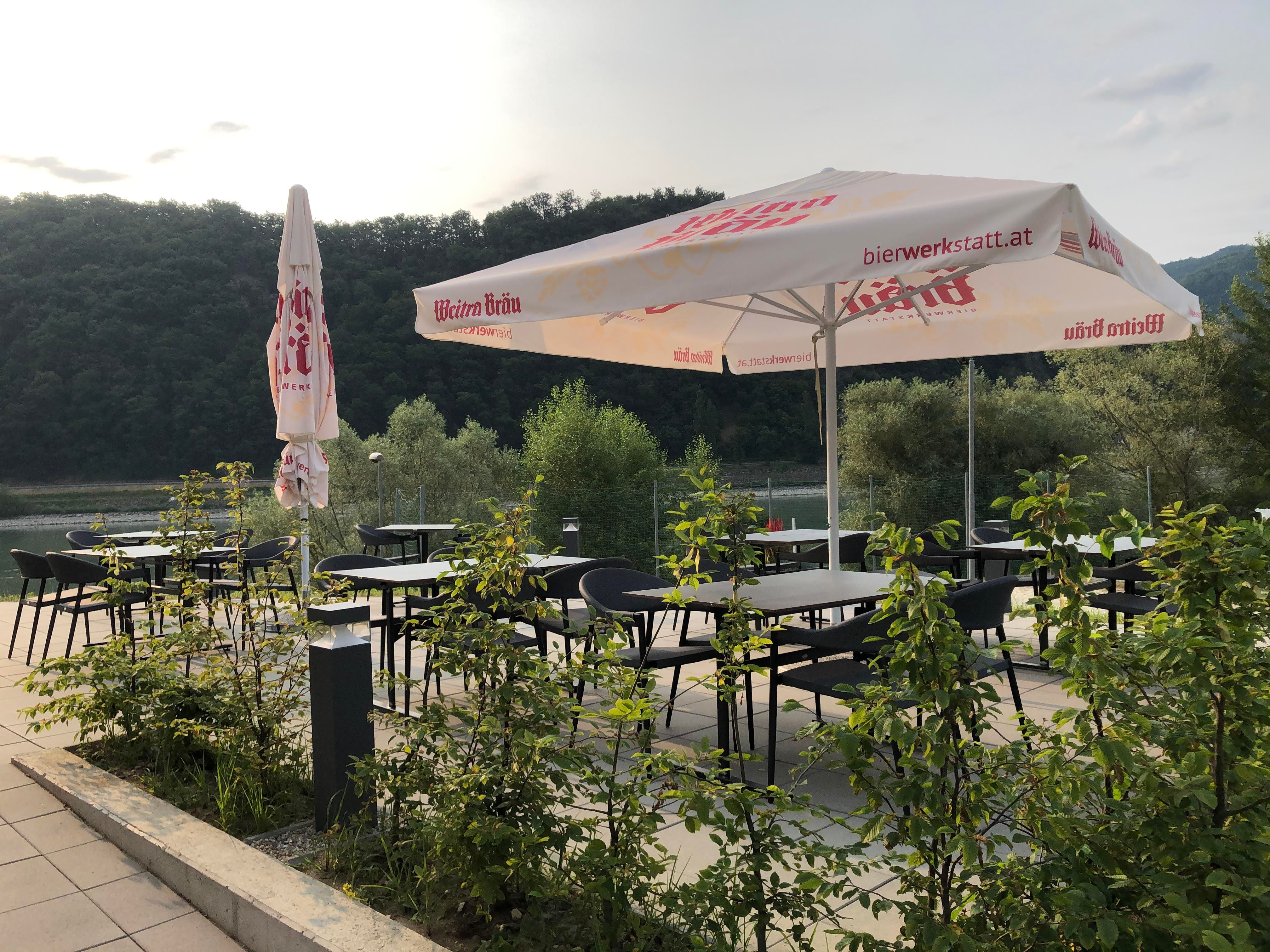 Terrasse mit Tischen, Stühlen und Sonnenschirmen, umgeben von Natur.