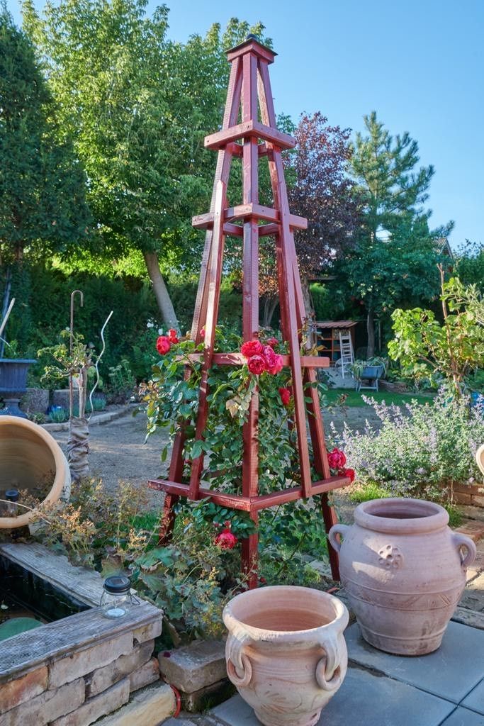 Ein Garten mit einer roten Rankhilfe, an der Rosen wachsen, umgeben von Töpfen und Pflanzen.