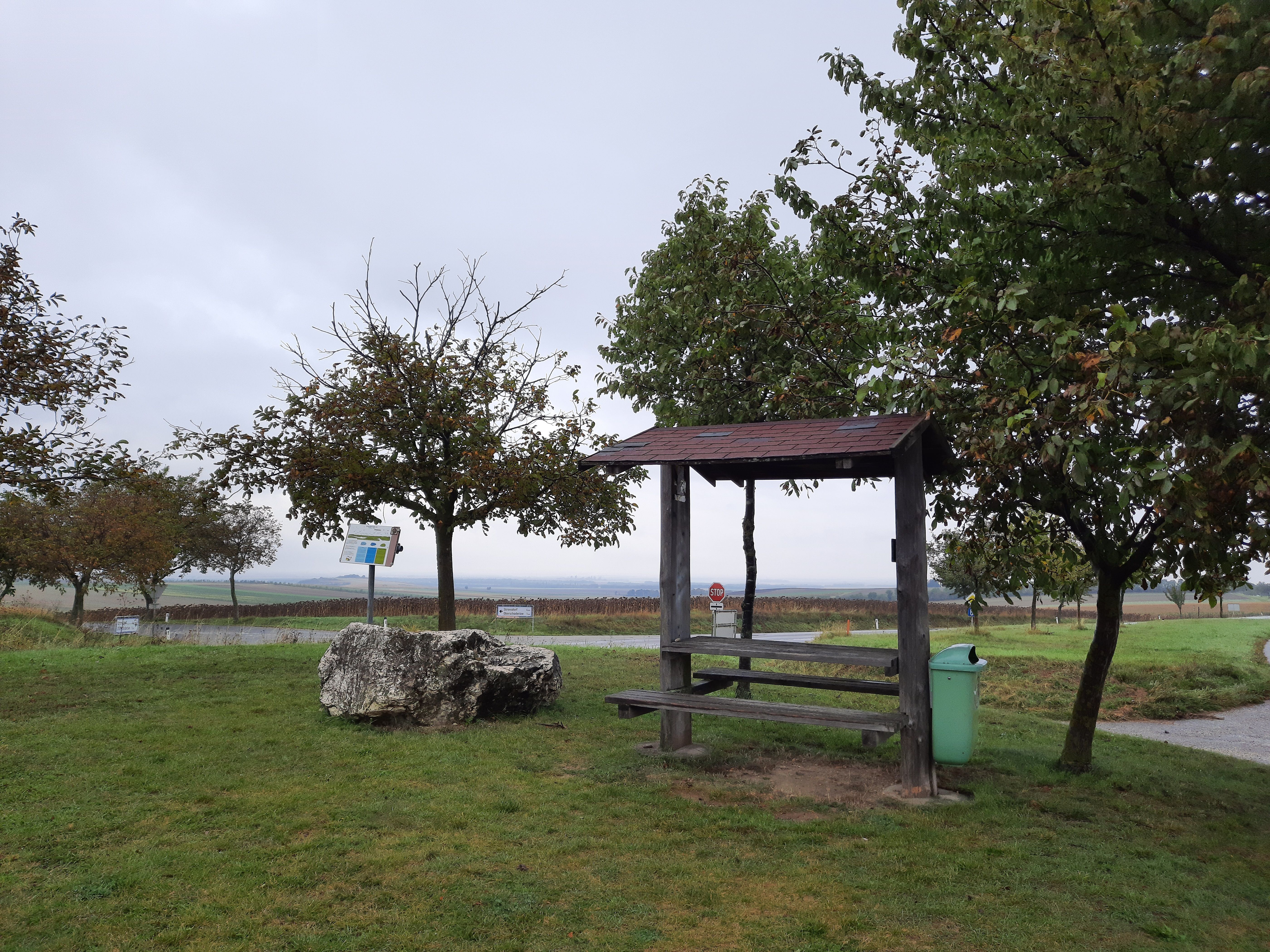 Ein Rastplatz mit Holzbänken und einem Tisch, umgeben von Bäumen und einer Wiese, unter einem bewölkten Himmel.