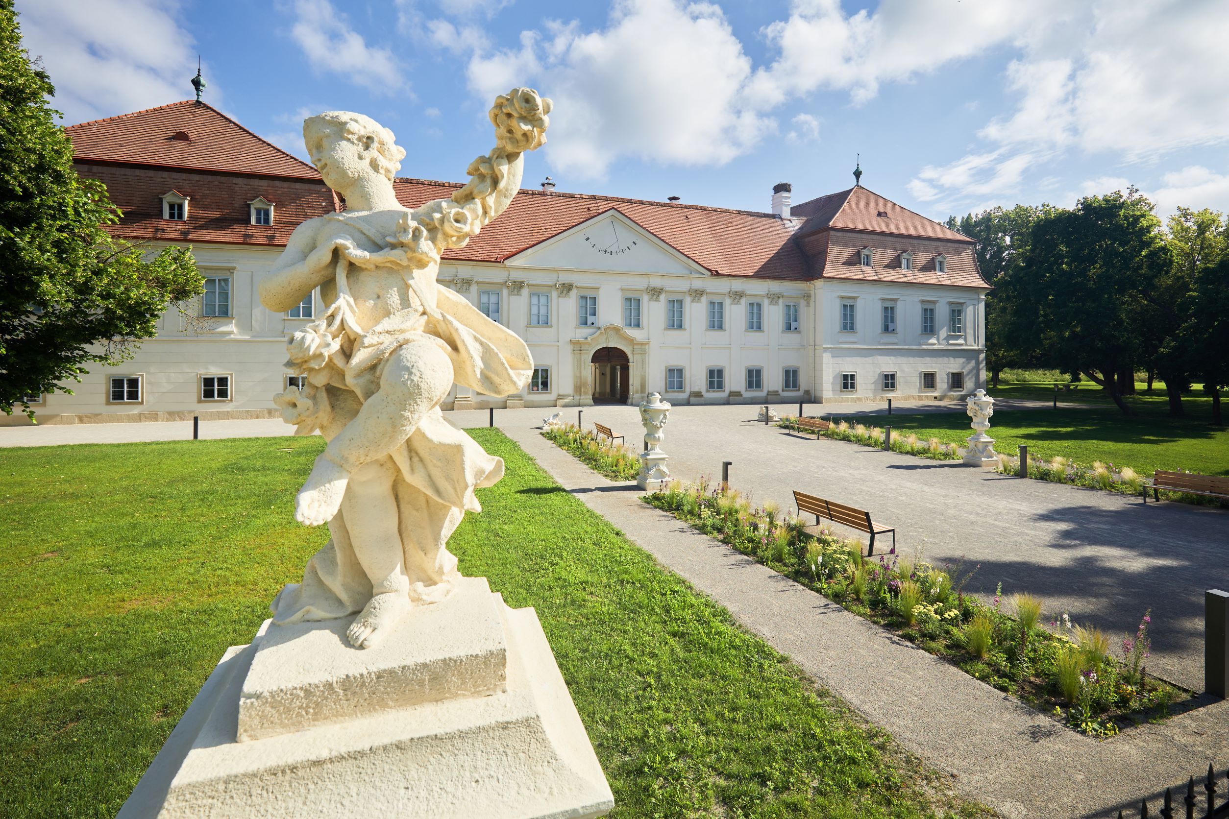 Barocke Statue vor einem historischen Gebäude mit rotem Dach und gepflegtem Garten.