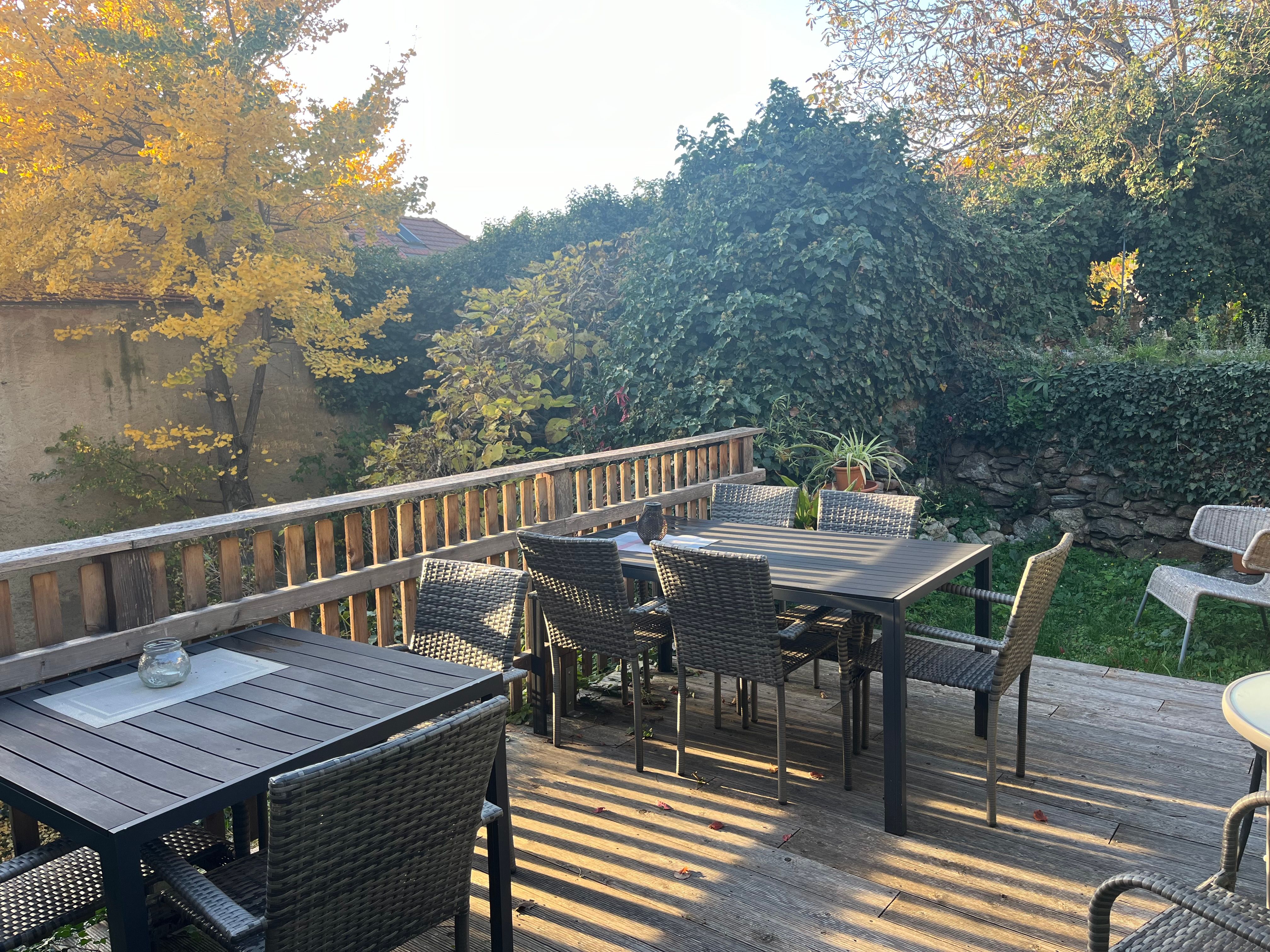 Terrasse mit Holztischen und Stühlen, umgeben von herbstlichen Bäumen und Pflanzen.